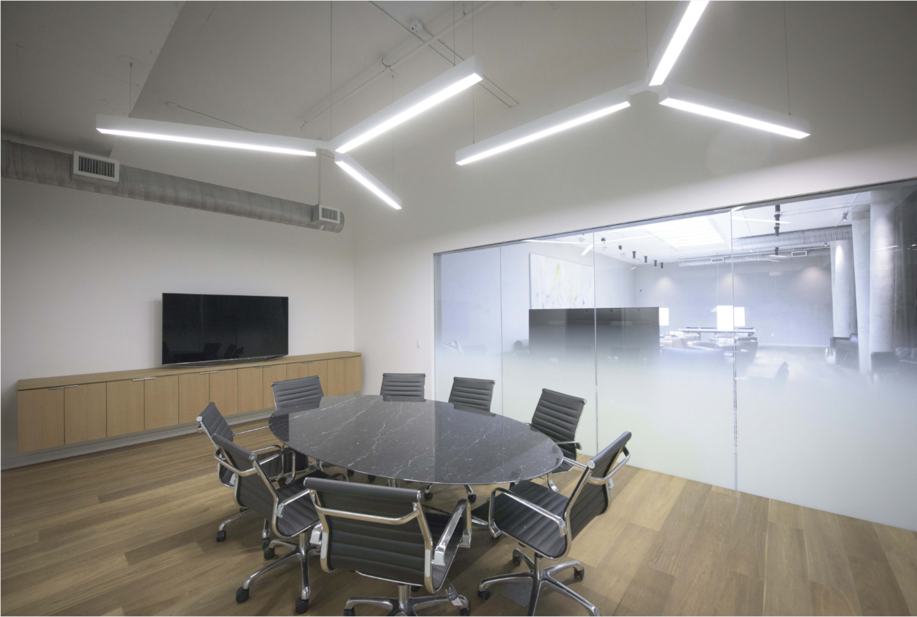 A modern conference room features a round marble table surrounded by sleek chairs, with linear overhead lighting and a glass wall leading to another workspace.