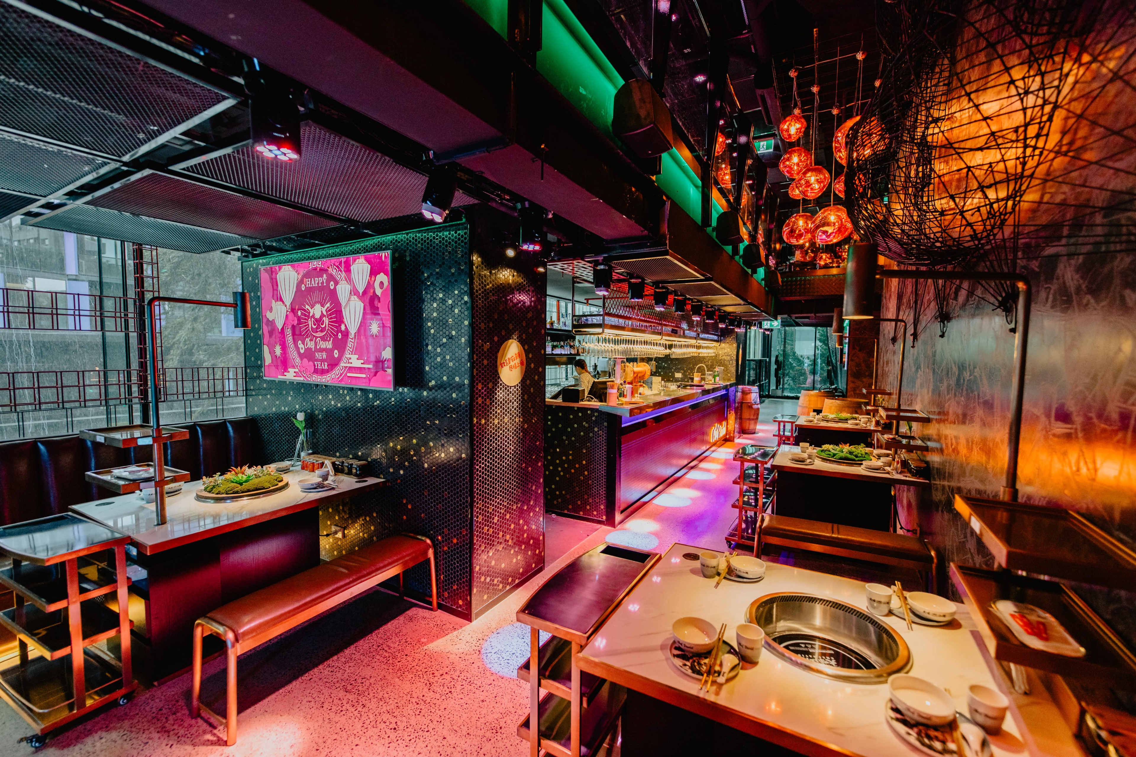 A modern restaurant interior with vibrant lighting, sleek seating arrangements, and a central cooking area featuring a grill at each table.