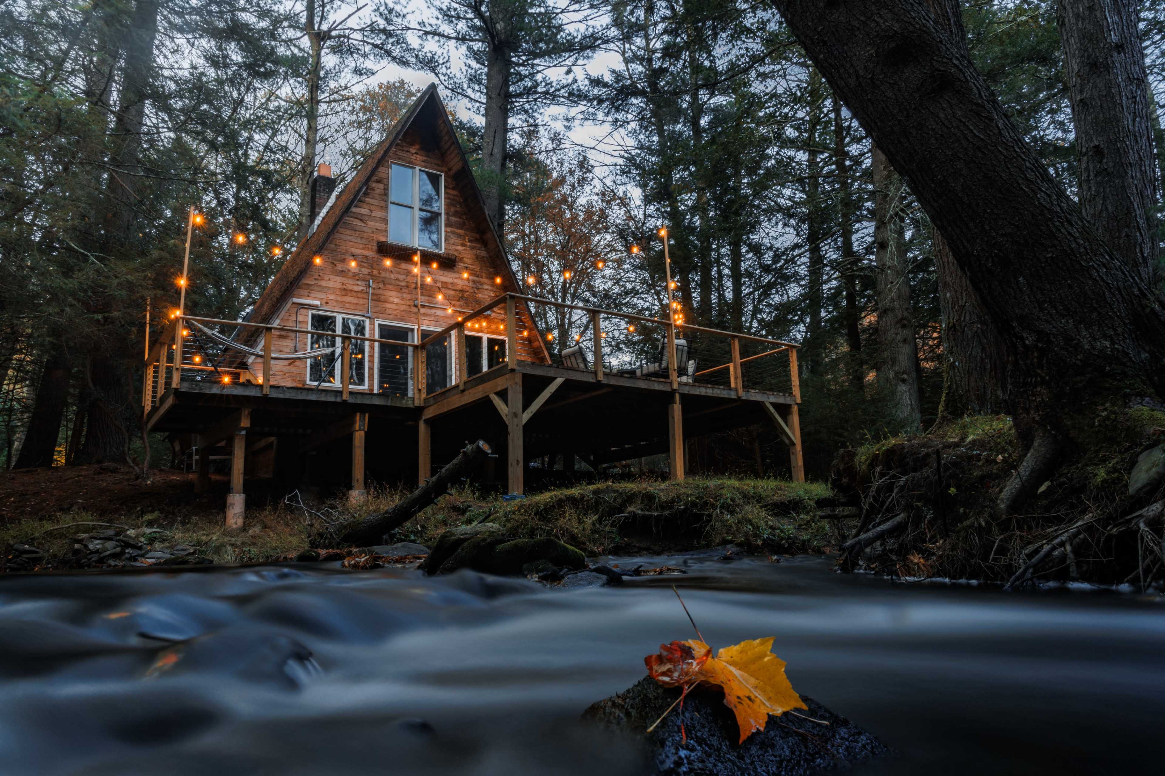 A wooden A-frame cabin with string lights sits on stilts by a flowing stream, surrounded by trees in a twilight setting.