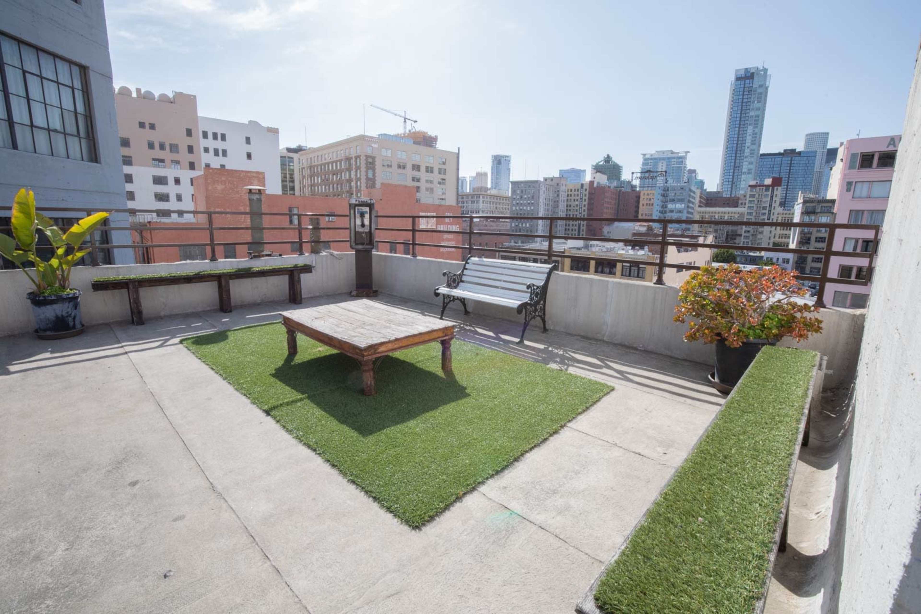 A rooftop terrace featuring a green carpeted area with a wooden coffee table, a bench, potted plants, and a city skyline in the background.