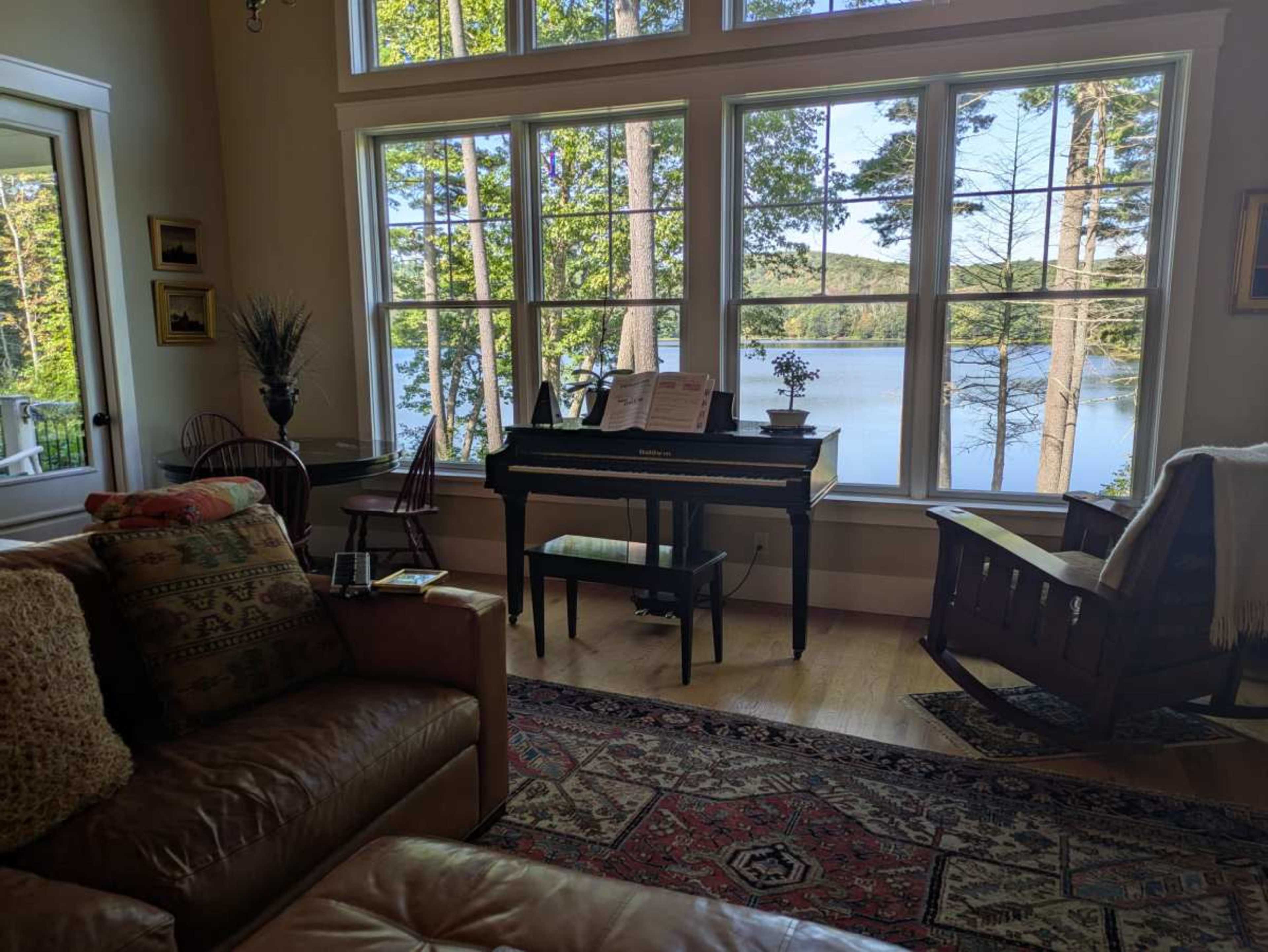 A cozy living room features a piano beside large windows overlooking a serene lake framed by trees.