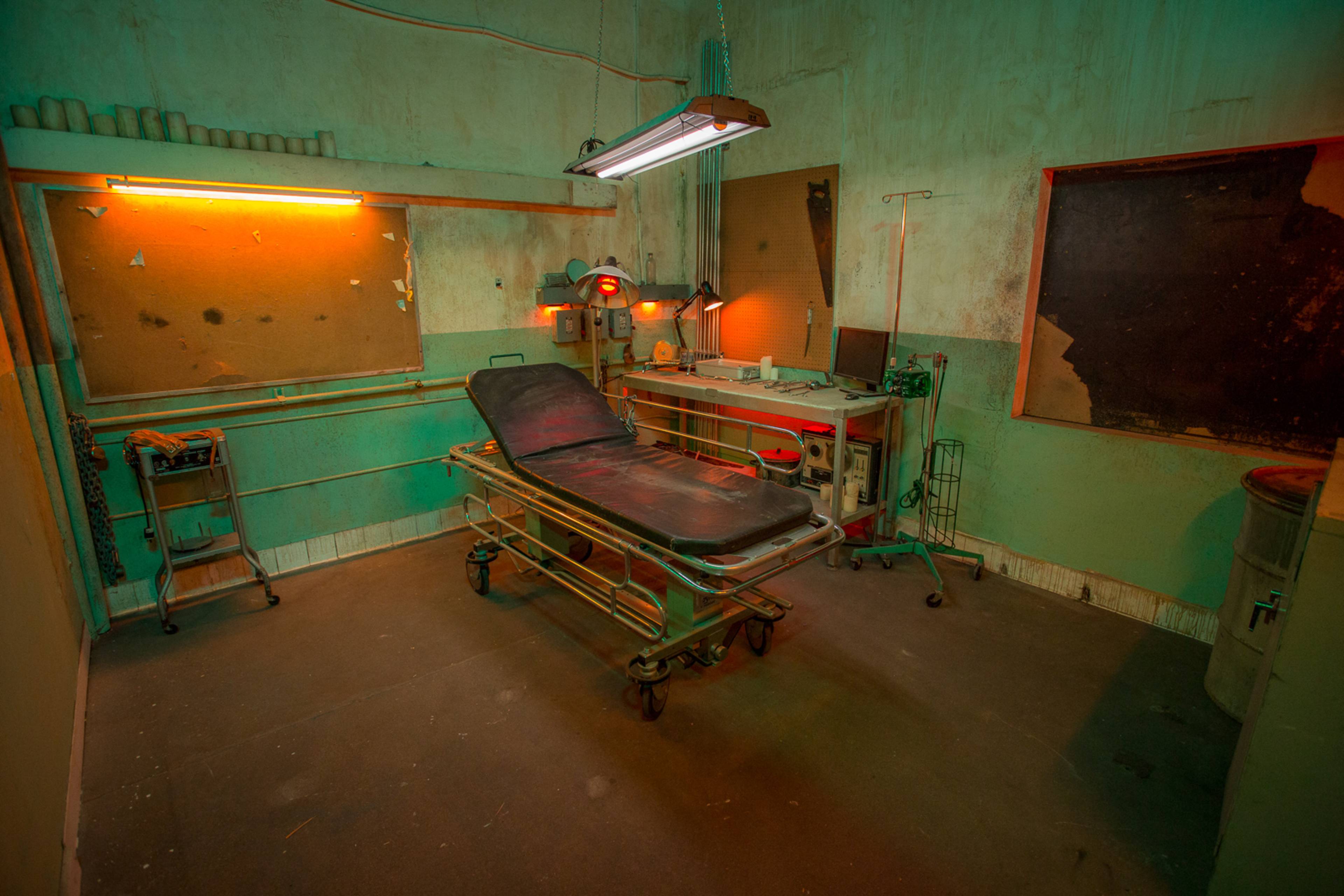 A dimly lit, empty examination room with a gurney, medical equipment, and weathered walls.