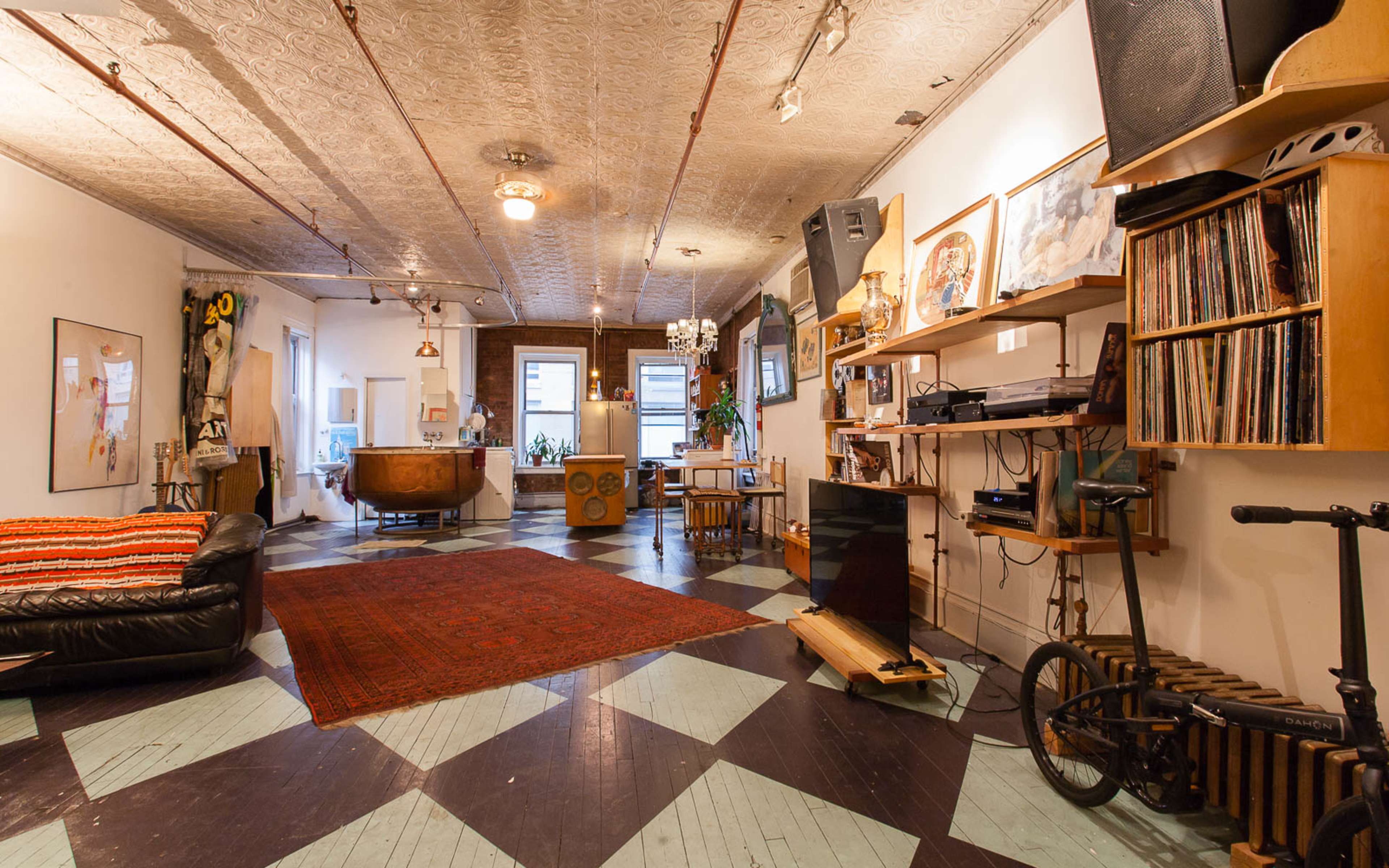 A spacious loft-style living area with a patterned floor, a black sofa, a wooden bar, a record player, and walls adorned with artwork.
