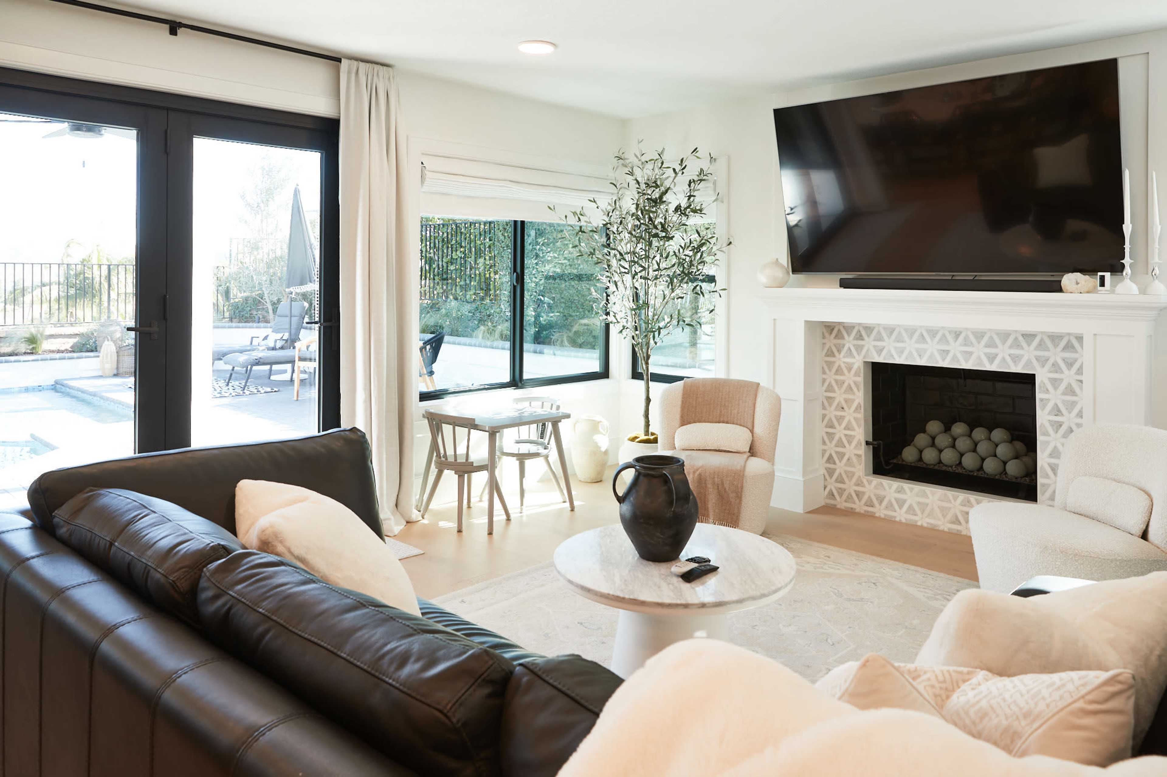 A modern living room with a black leather sofa, a round marble coffee table, and a fireplace, with large glass doors leading to an outdoor area with a pool.