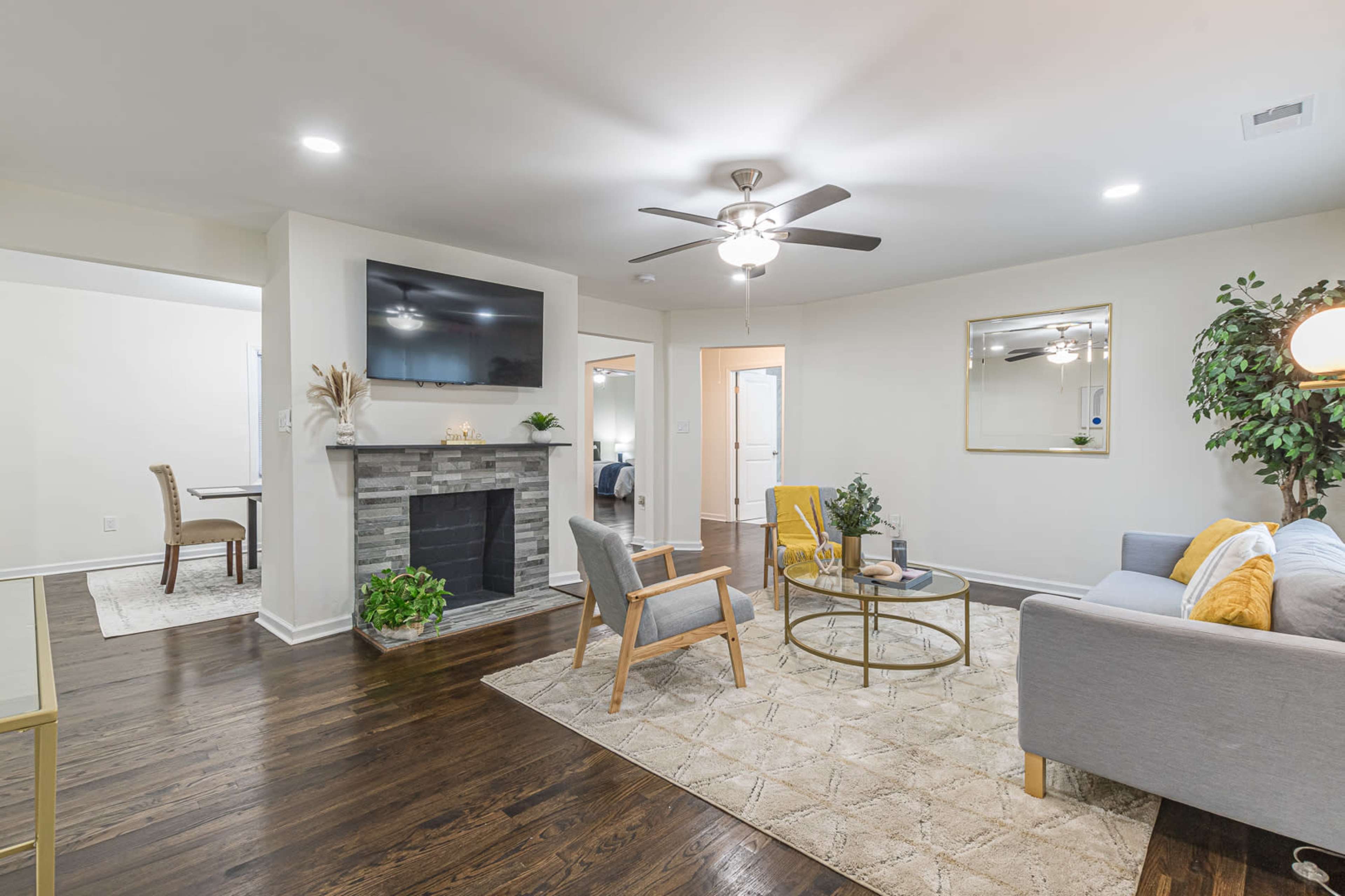 A modern living room features a gray sofa, a circular coffee table, a stone fireplace, and a large mirror, with a ceiling fan above and plants decorating the space.