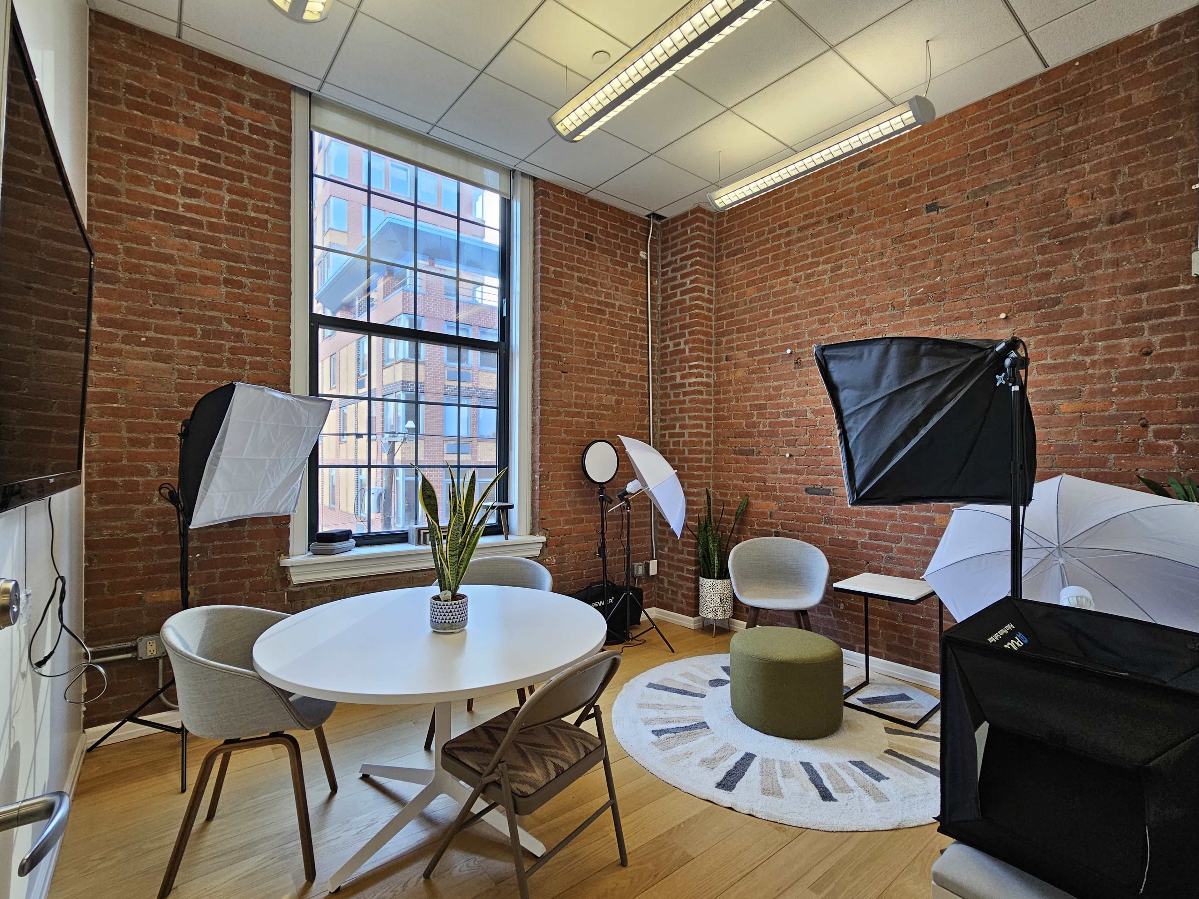 The image shows a small studio space with exposed brick walls, a round table, chairs, and several lighting setups for photography.