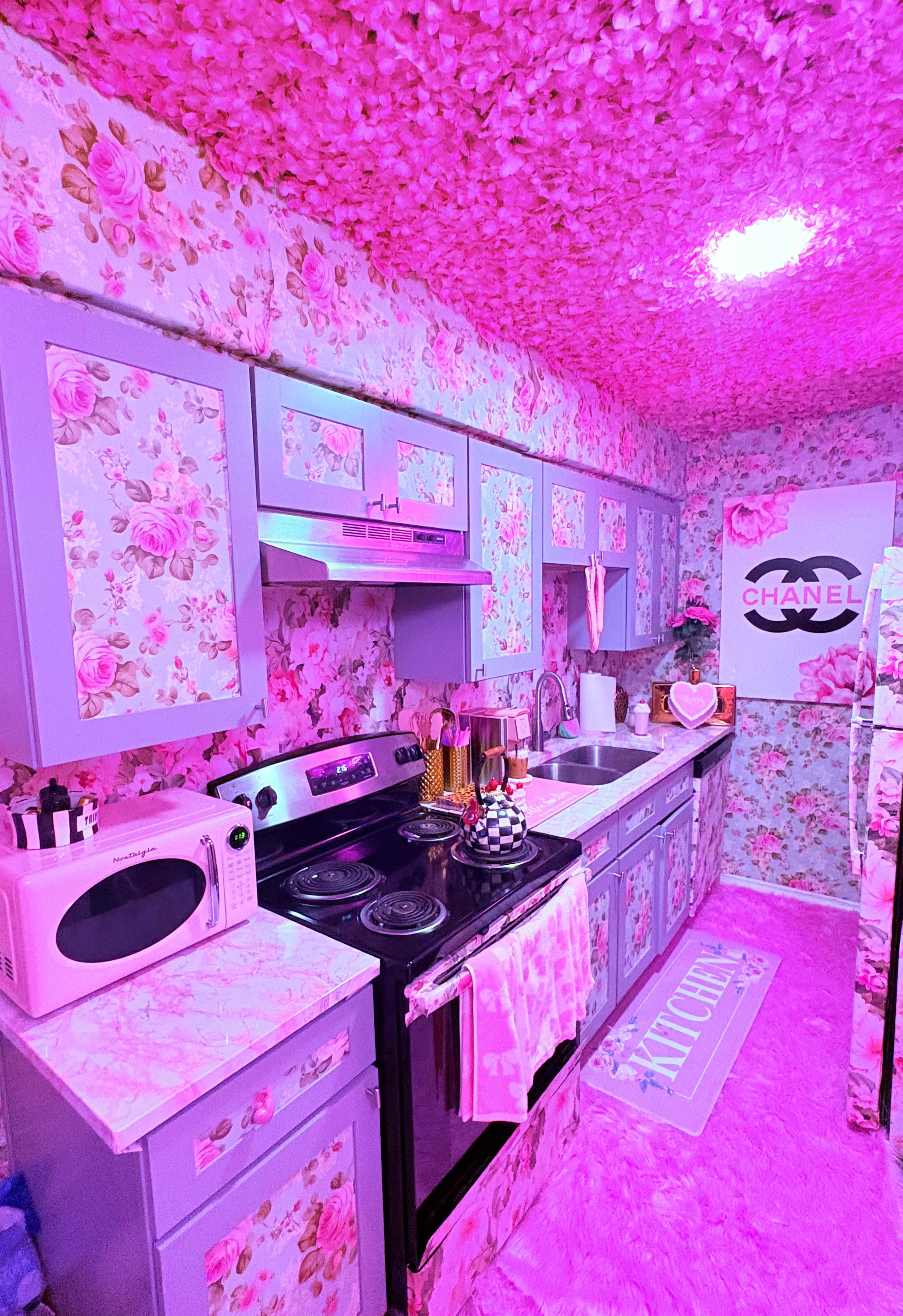 The image shows a kitchen with floral-patterned walls and ceiling, featuring pink decor, modern appliances, and a sign with the Chanel logo.