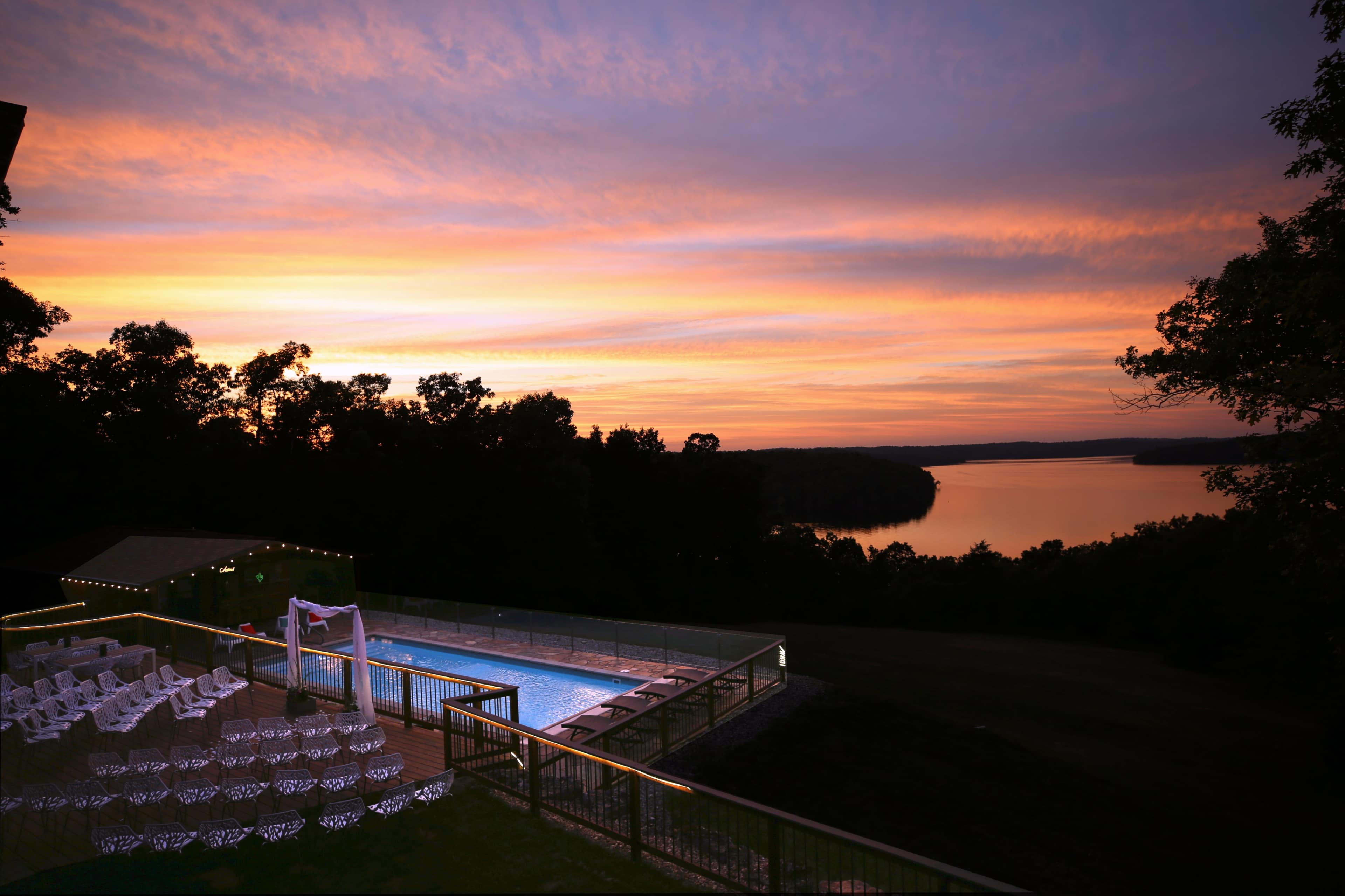 A swimming pool area with white chairs arranged around it, overlooking a lake at sunset.