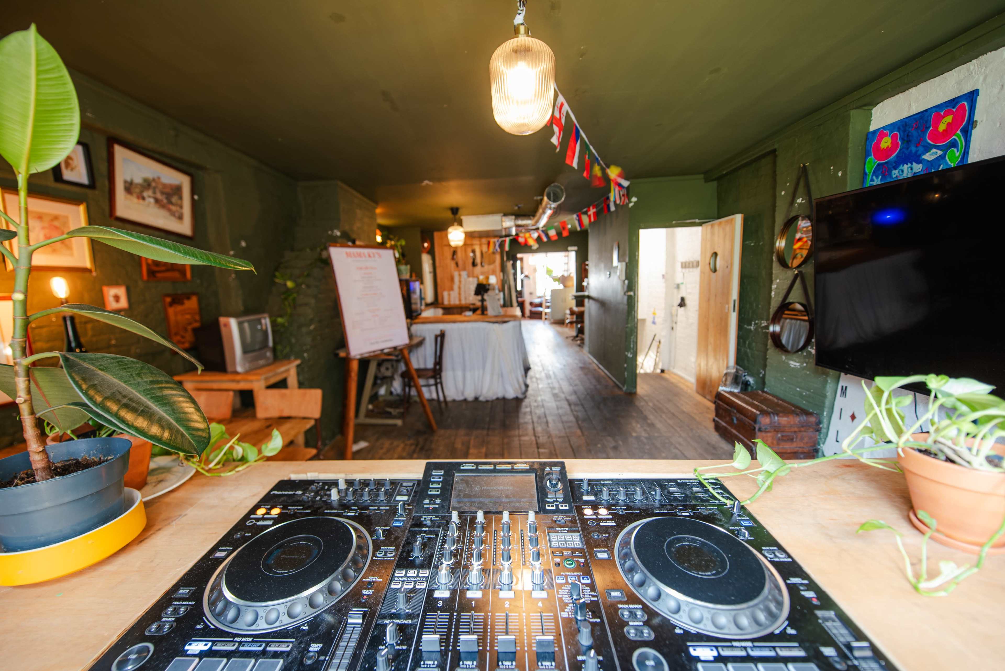 The image shows a DJ controller in the foreground with a vibrant café or bar setting in the background, featuring decorative flags and a cozy atmosphere.