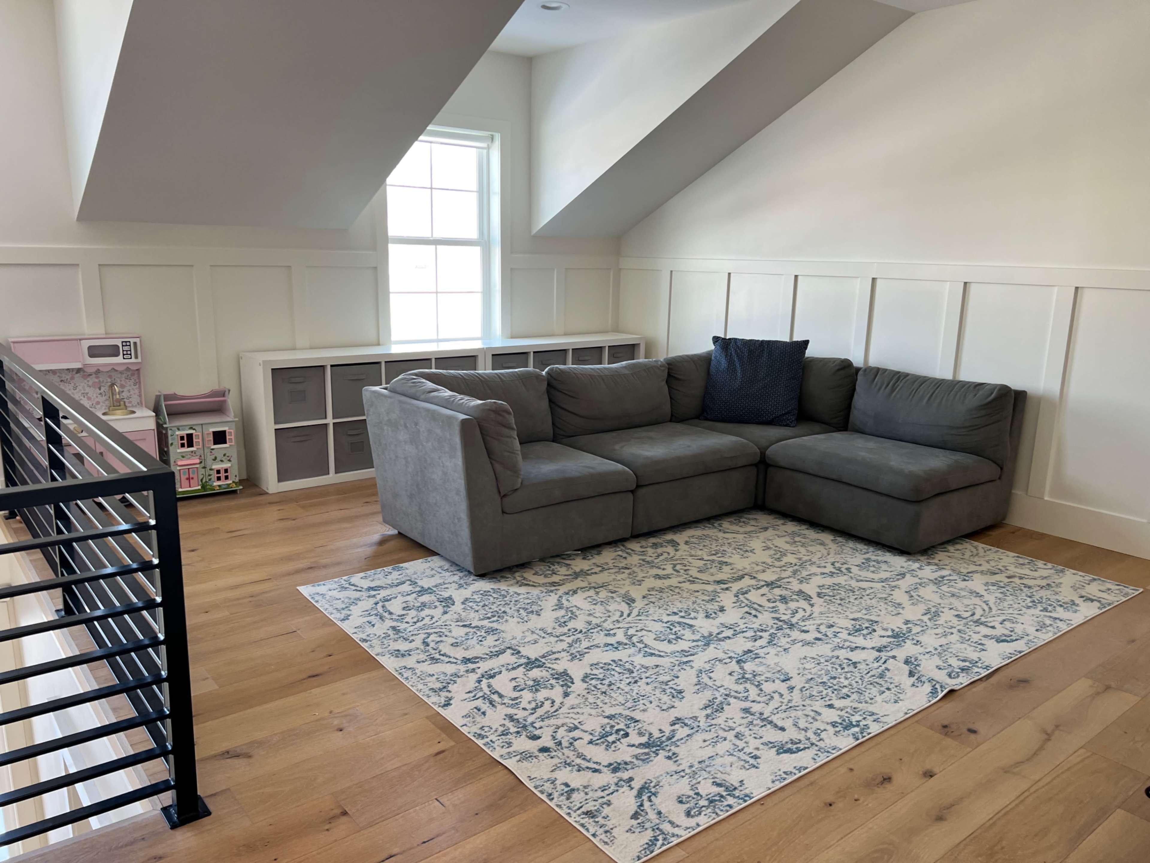 The image shows a well-lit room with a gray sectional couch, a blue and white patterned rug, and a children's play area featuring a toy kitchen and dolls.