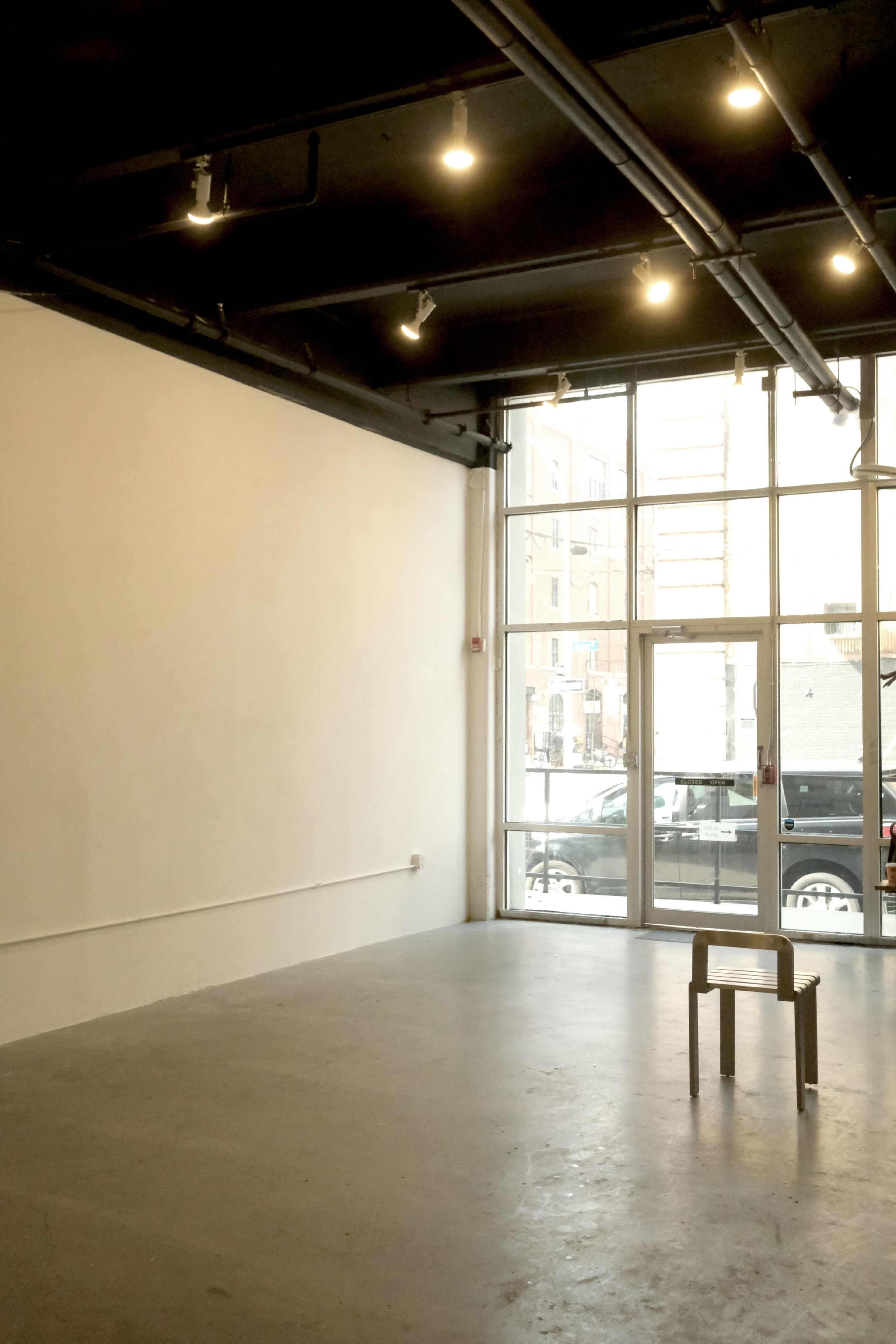 The image shows a minimalist interior of an art gallery or studio with a wooden chair positioned in the center, and large glass doors revealing a view of the street outside.