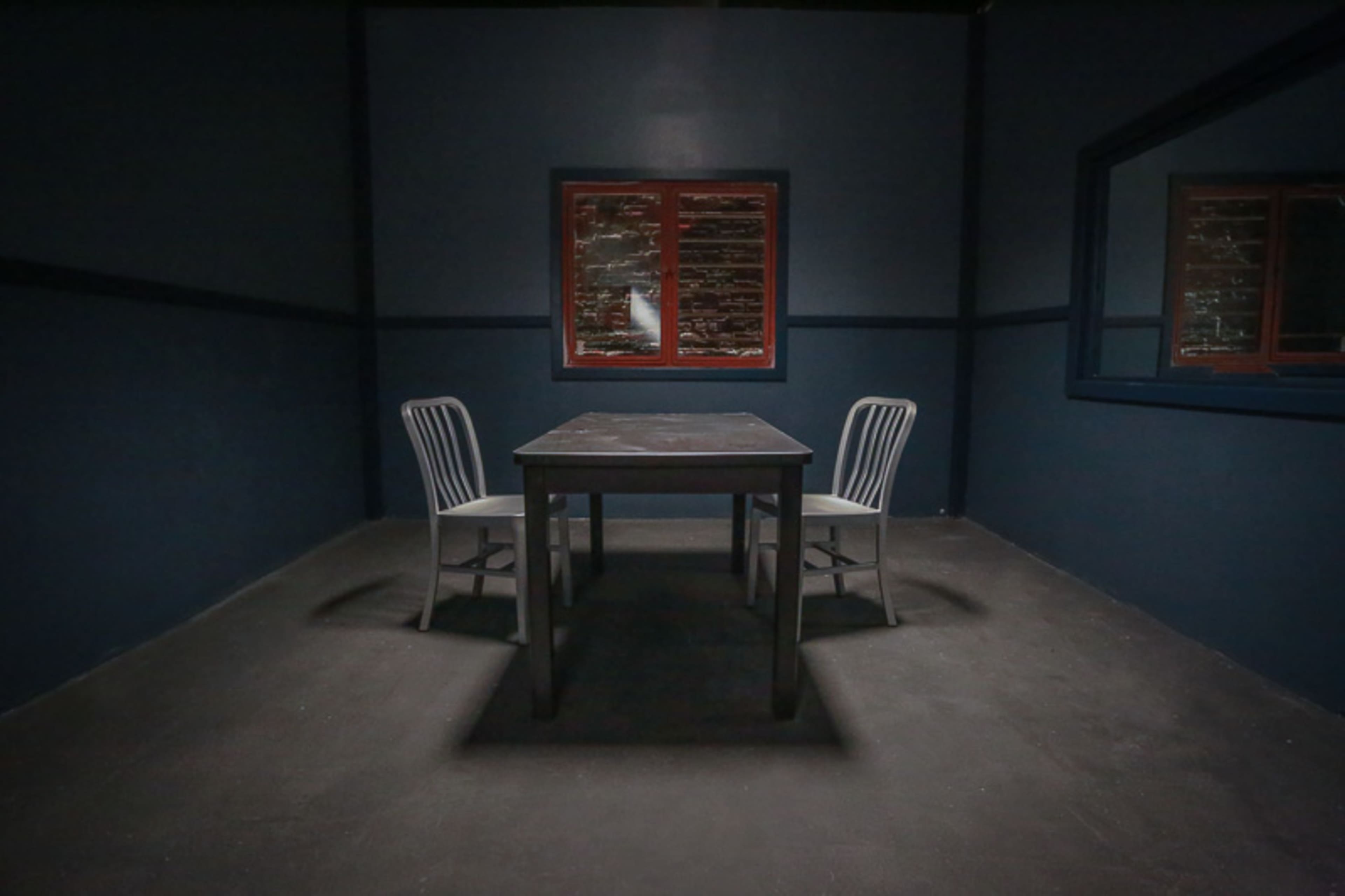 A dimly lit room features a table surrounded by two chairs and a window with a red frame.