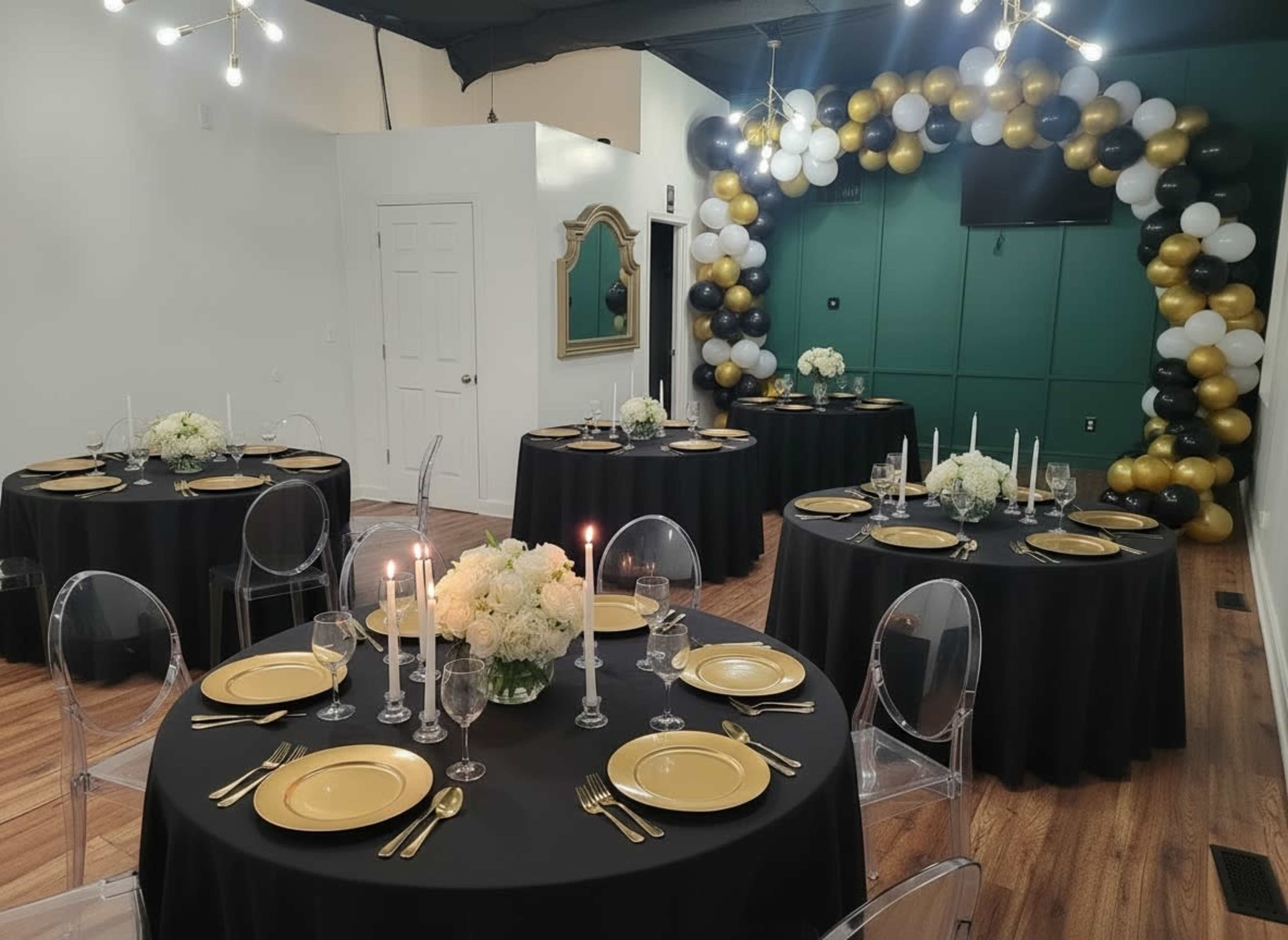 The image shows a decorated indoor event space with several round tables set for a formal gathering, featuring black tablecloths, gold plates, and floral centerpieces, alongside a balloon arch in black, gold, and white.