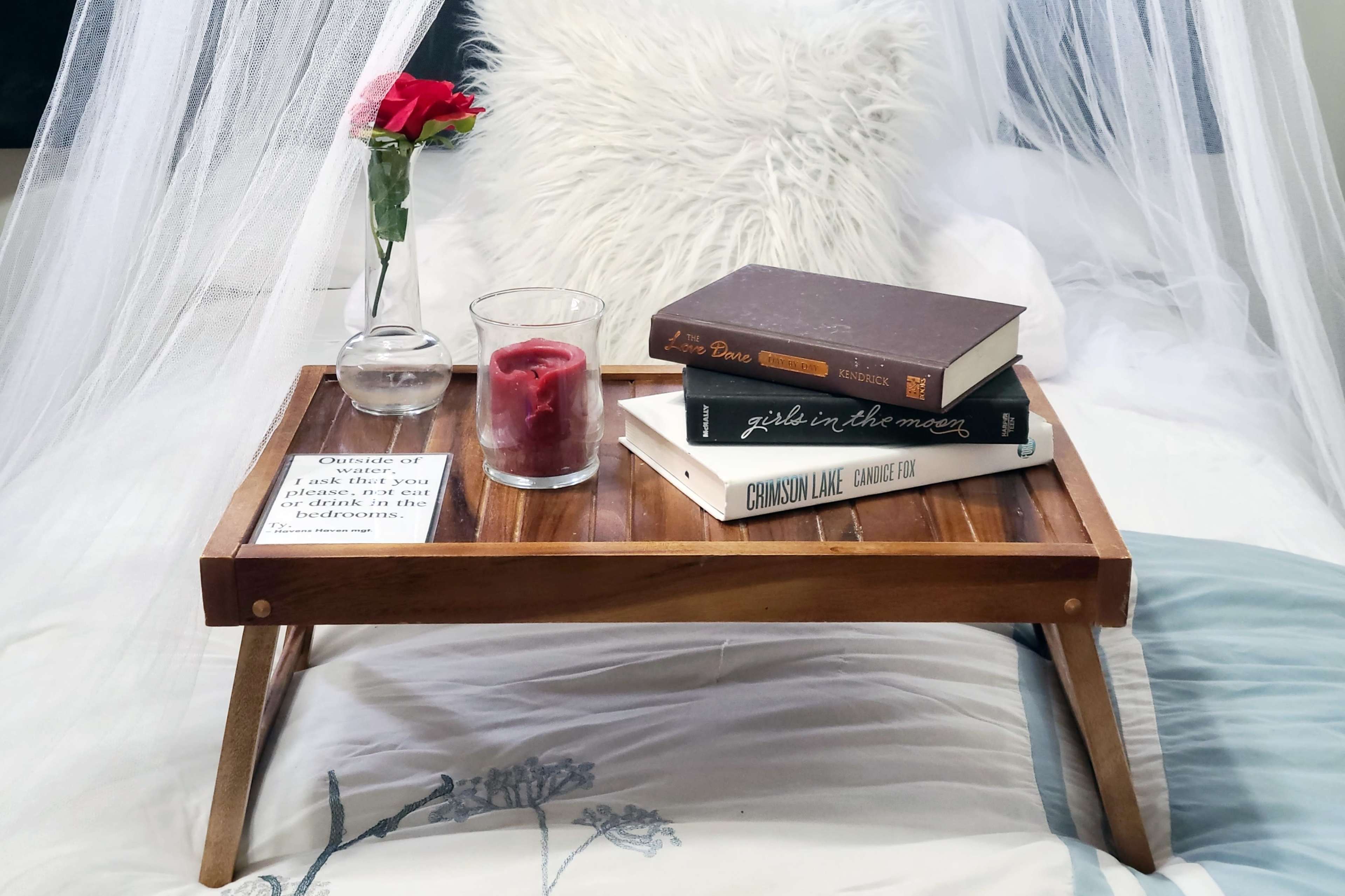A wooden breakfast tray with a rose, a glass of water, a candle, and three books sits on a bed draped in a light canopy.