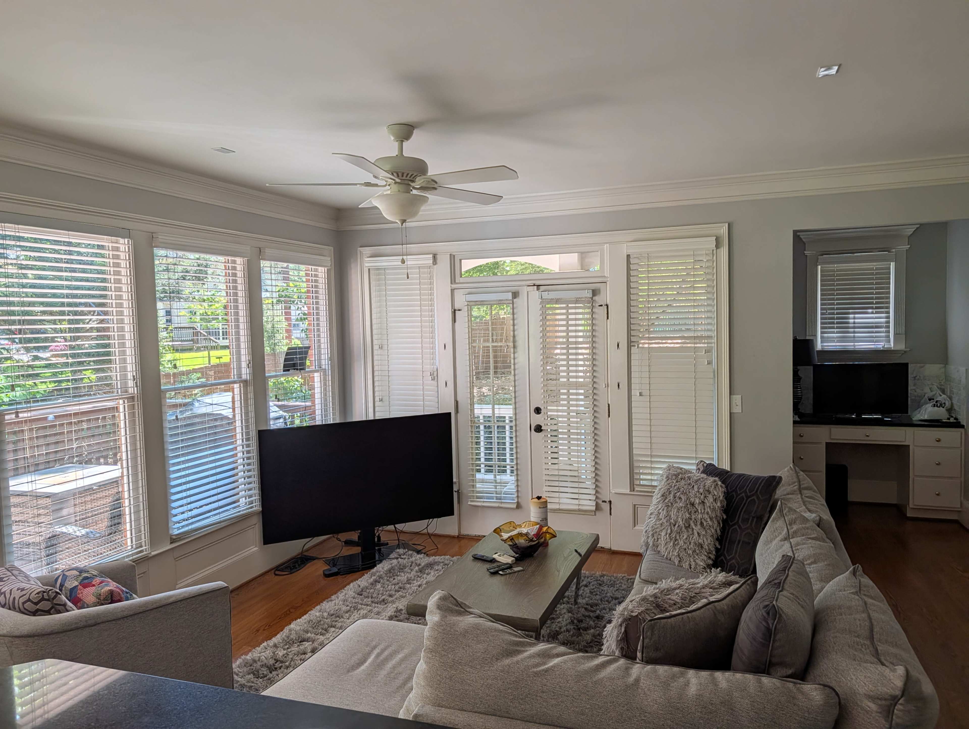 The image shows a living room with a large sectional sofa, a coffee table, a television, and windows with plantation shutters letting in natural light.