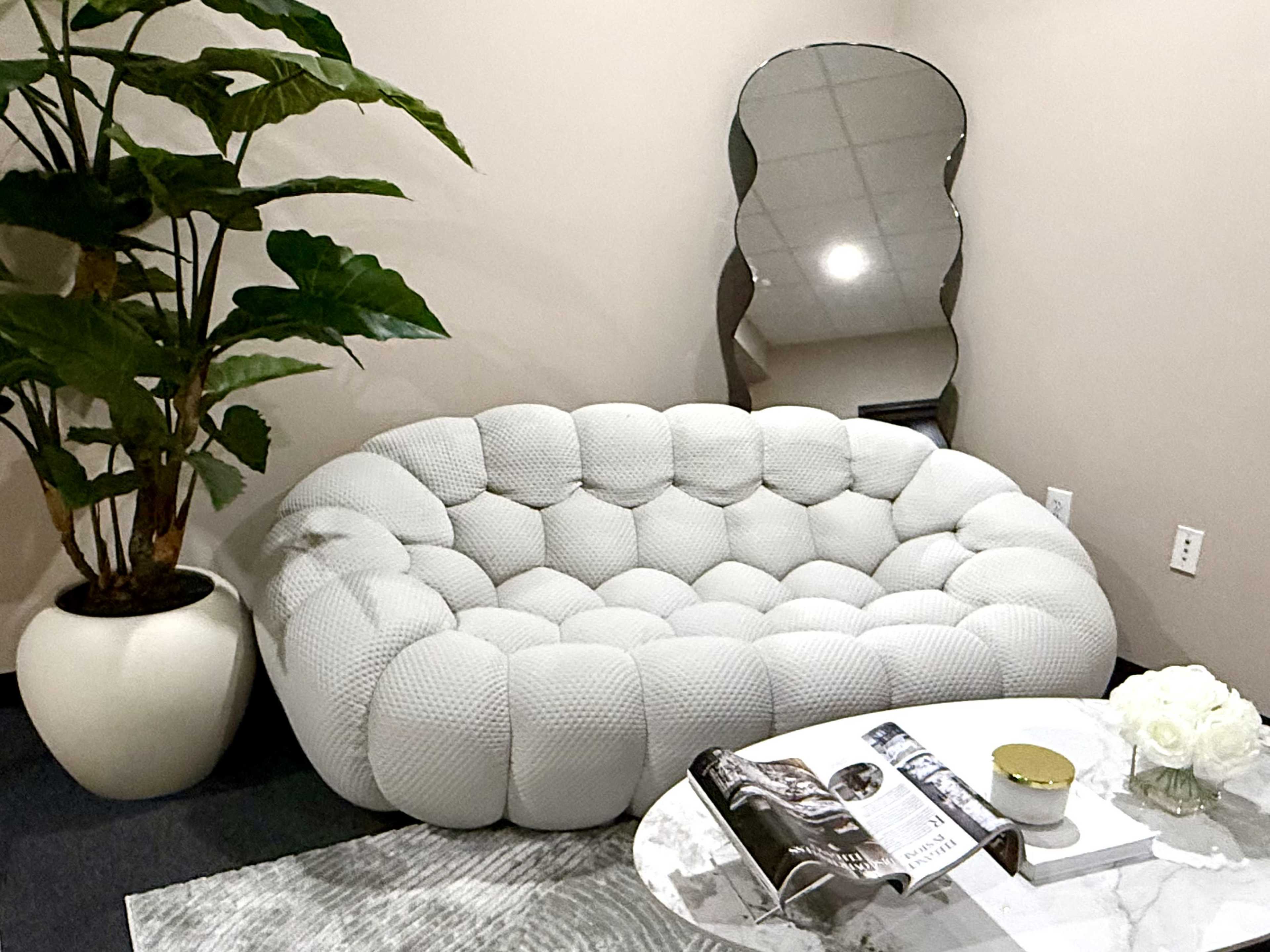 The image features a white cushioned sofa arranged next to a large potted plant and a mirrored wall decoration, complemented by a round coffee table with a magazine and decorative items.