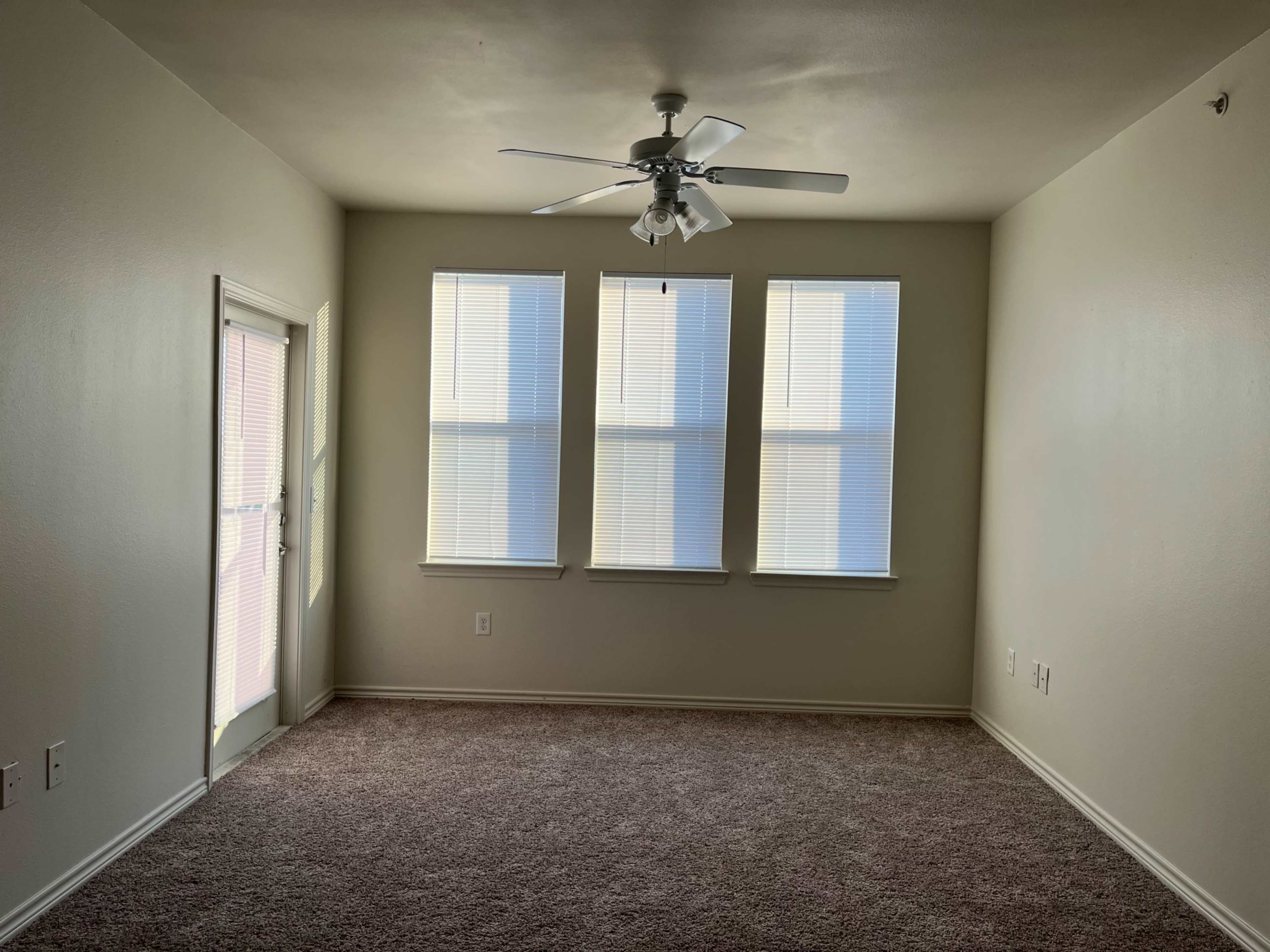 A vacant room with three windows, a ceiling fan, and a door leading outside.