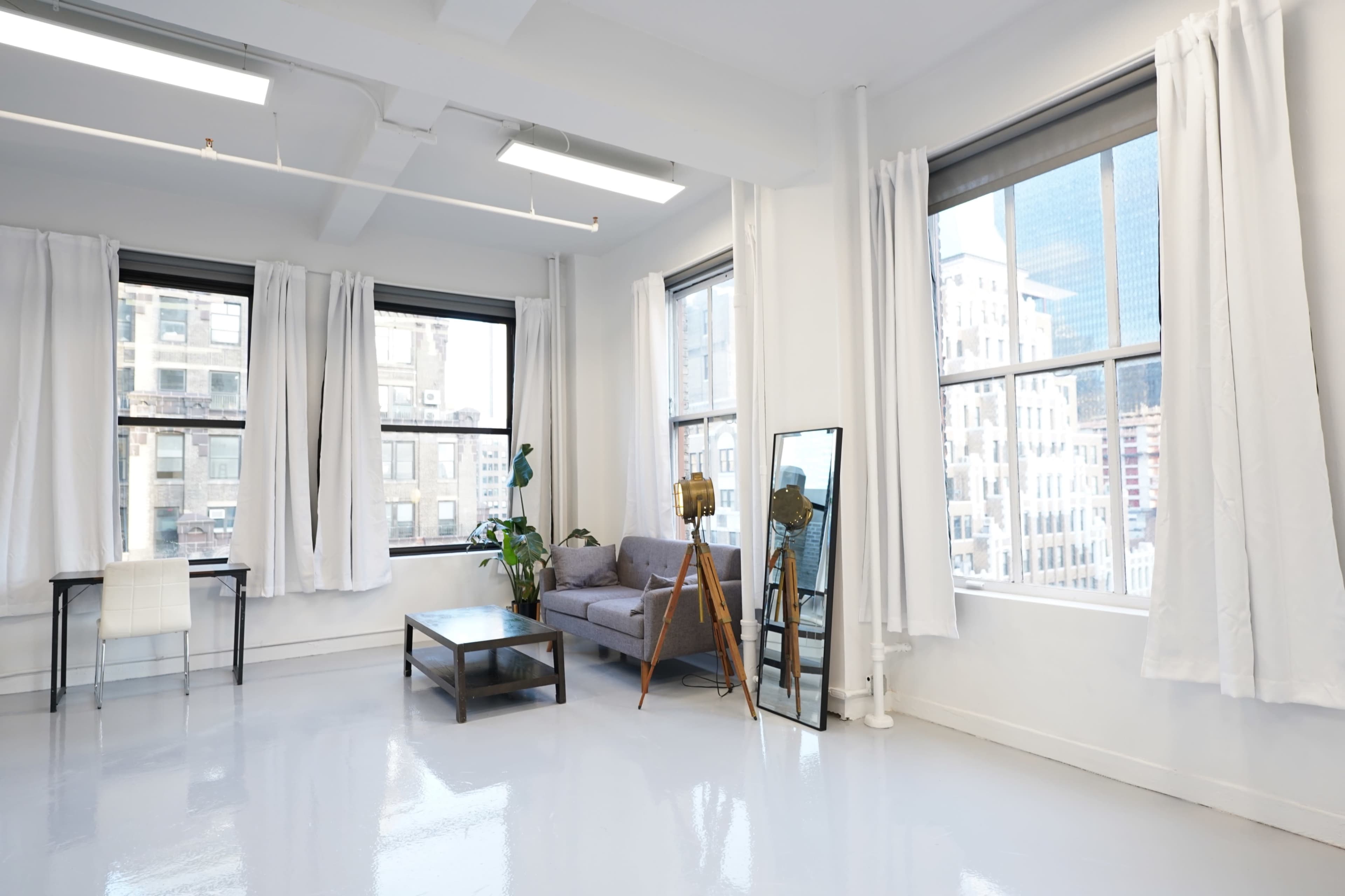A bright, modern room features large windows, white walls, a gray couch, a wooden coffee table, and a floor mirror.