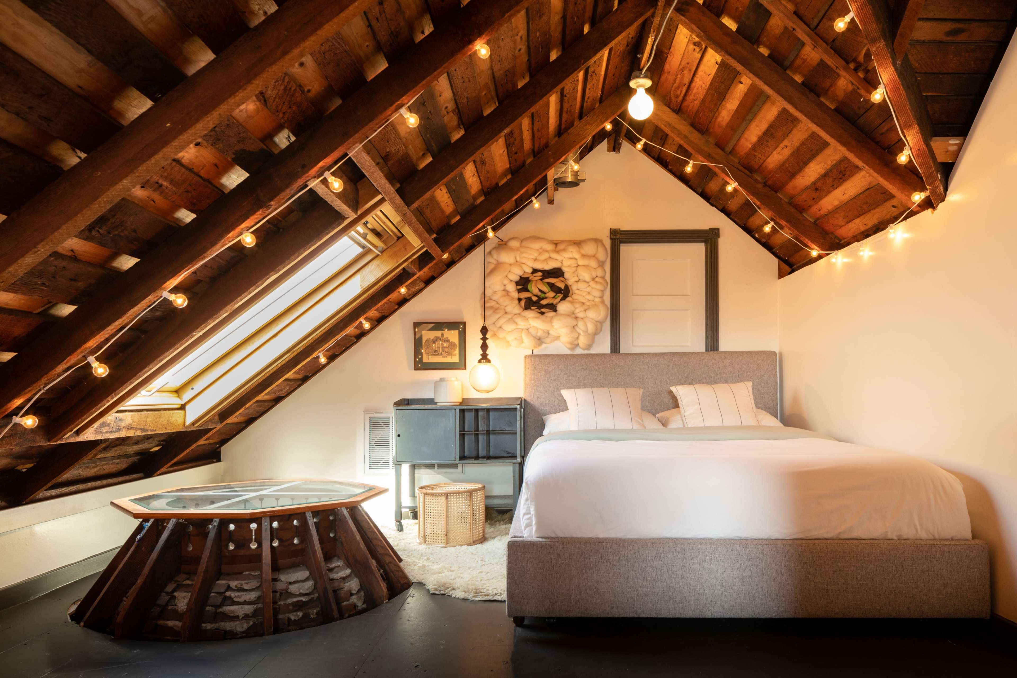 The cozy bedroom features a sloped wooden ceiling with hanging lights, a bed with gray upholstery, and a circular coffee table made of wood and stone.
