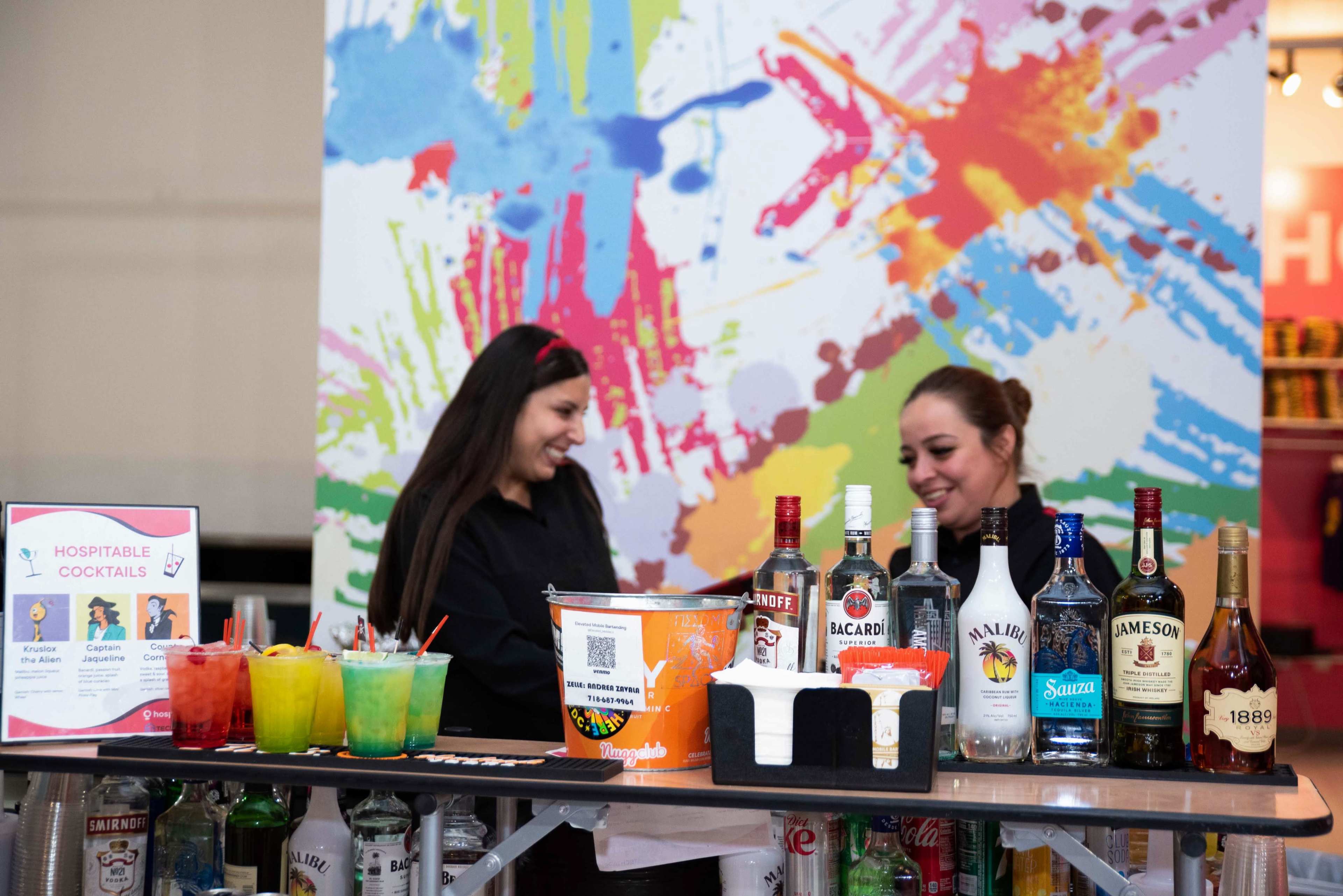 Two women stand behind a bar, interacting while surrounded by colorful splatter art and a variety of liquor bottles.
