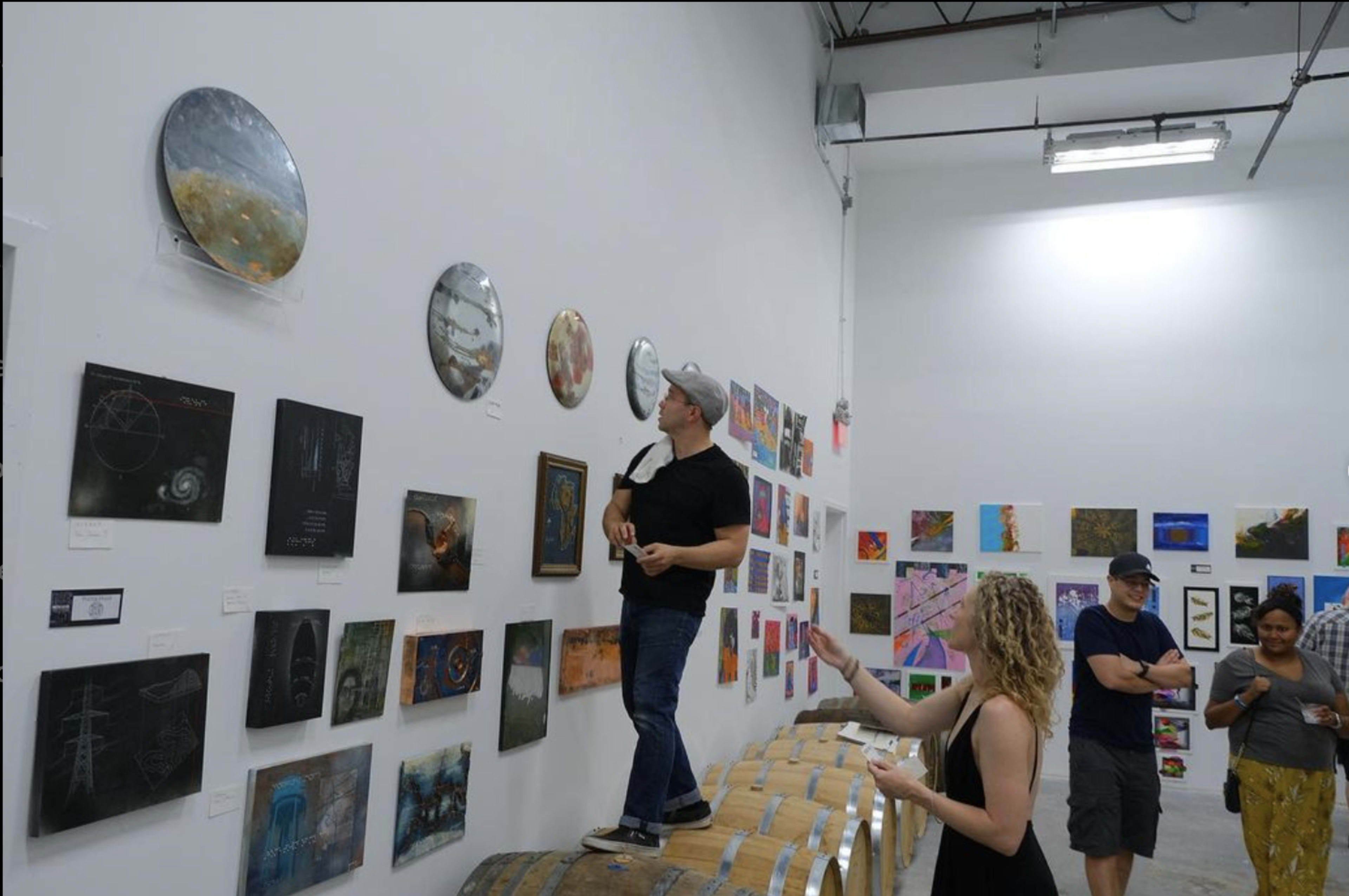 A man stands on a barrel while inspecting art pieces on a wall in a gallery, as three other individuals observe nearby.