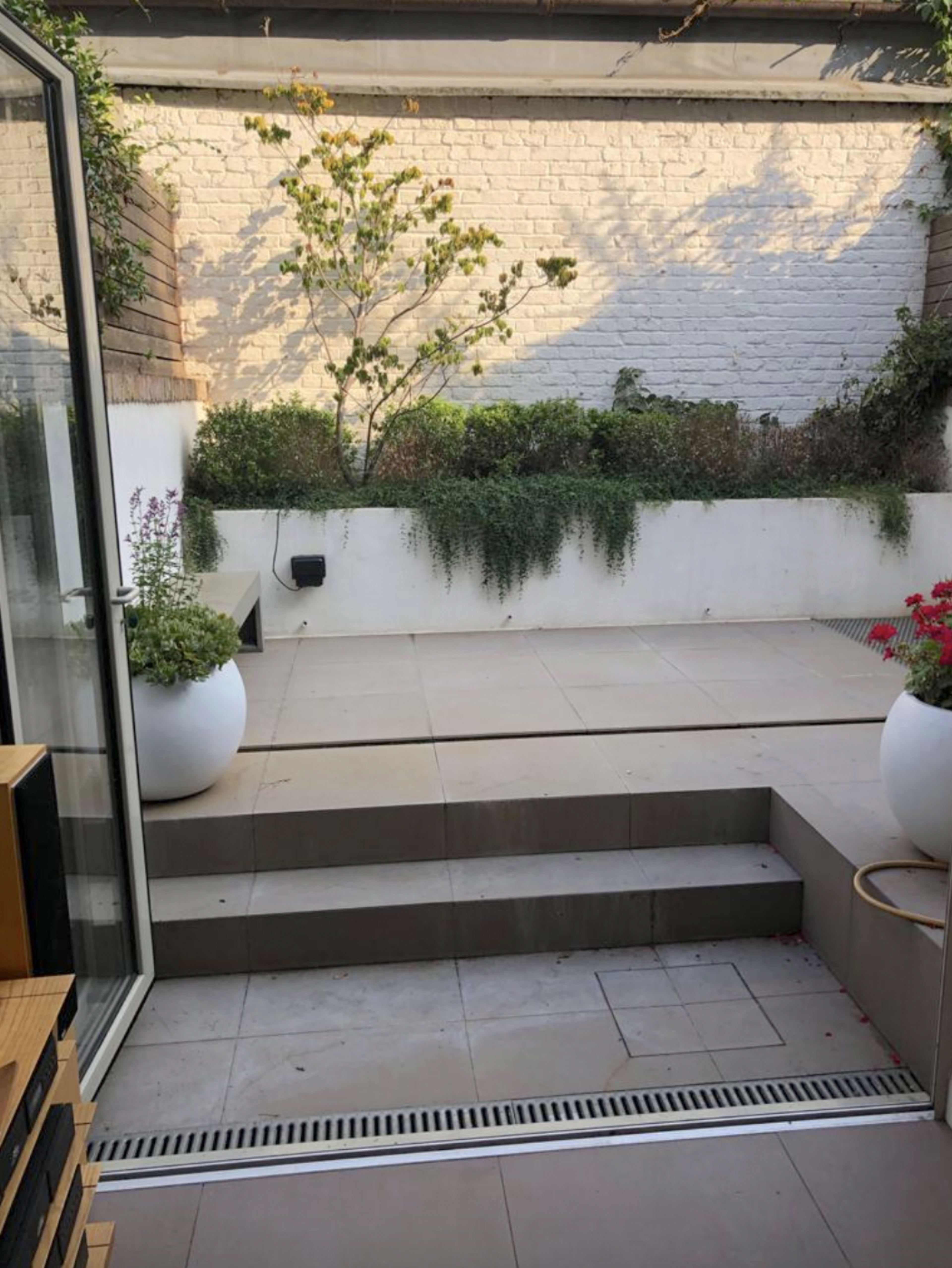 A courtyard with a paved area, steps leading down, and potted plants against a white wall.