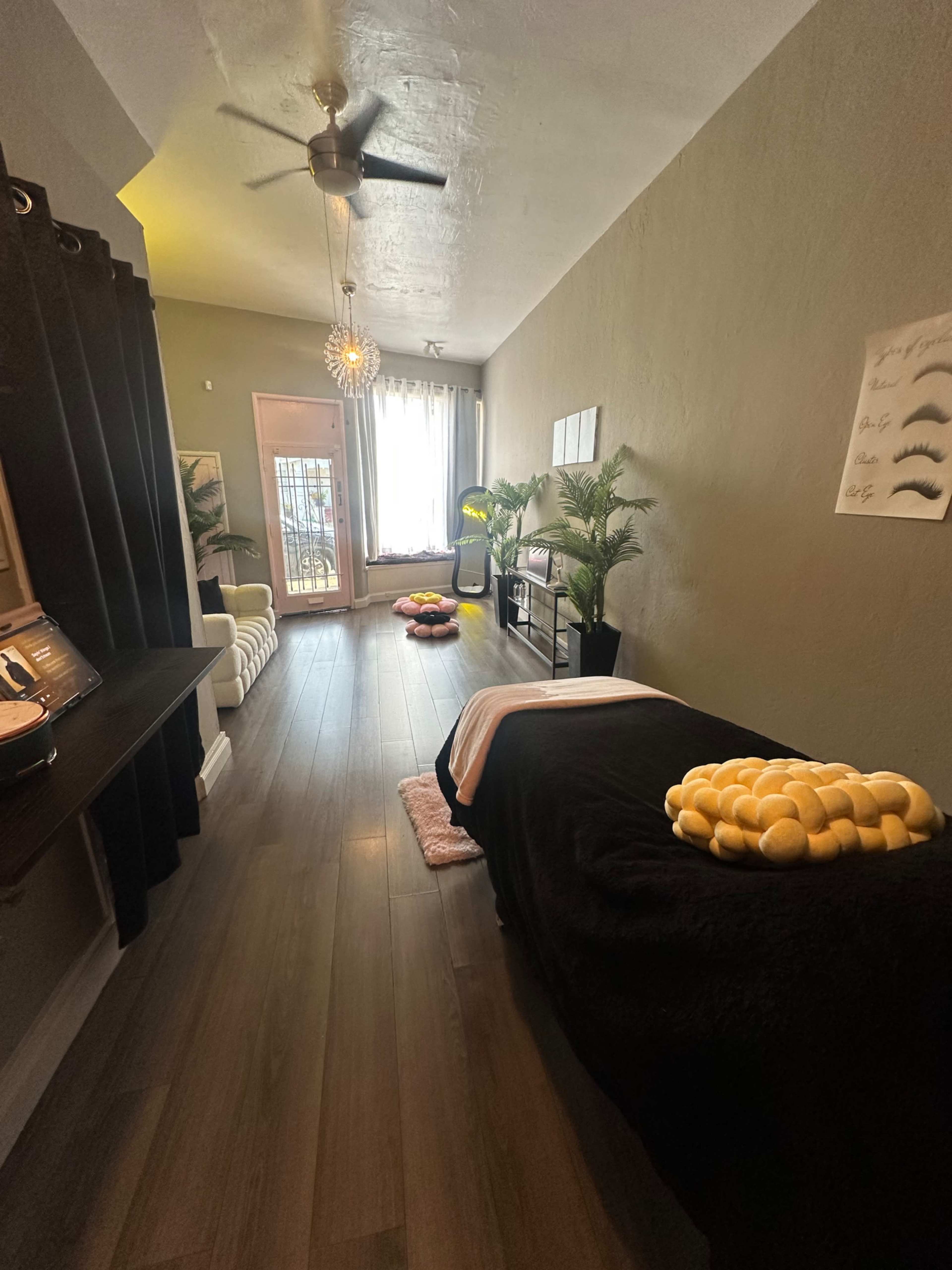 A spacious room features a massage table with a black cover, surrounded by potted plants and natural light from a large window.