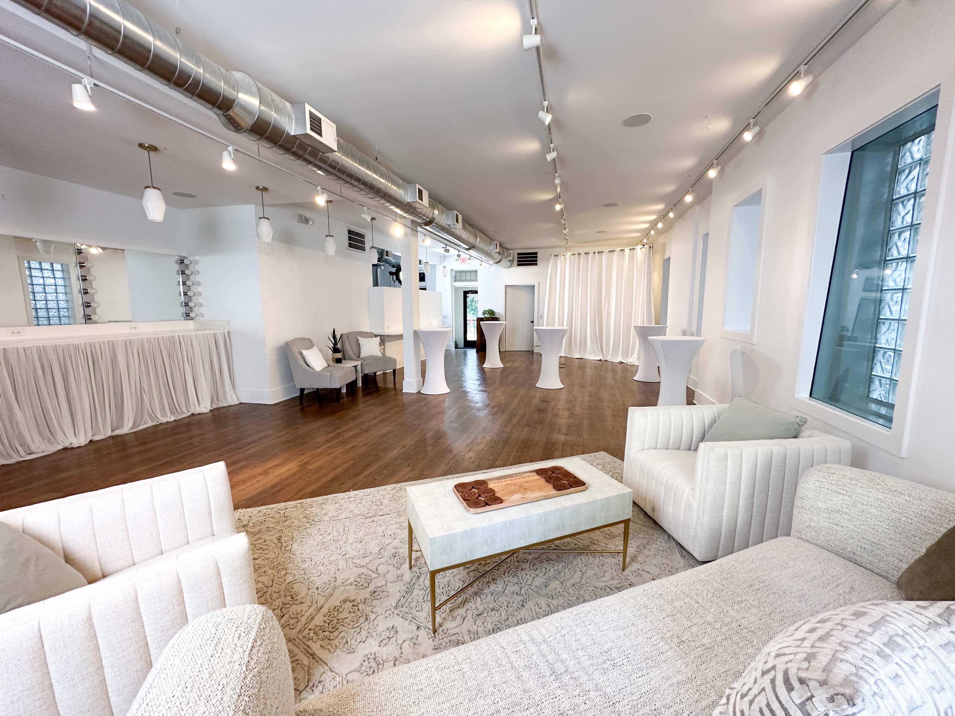 The image shows a spacious, well-lit event space featuring white walls, wooden flooring, and several lounge and cocktail tables arranged for gatherings.