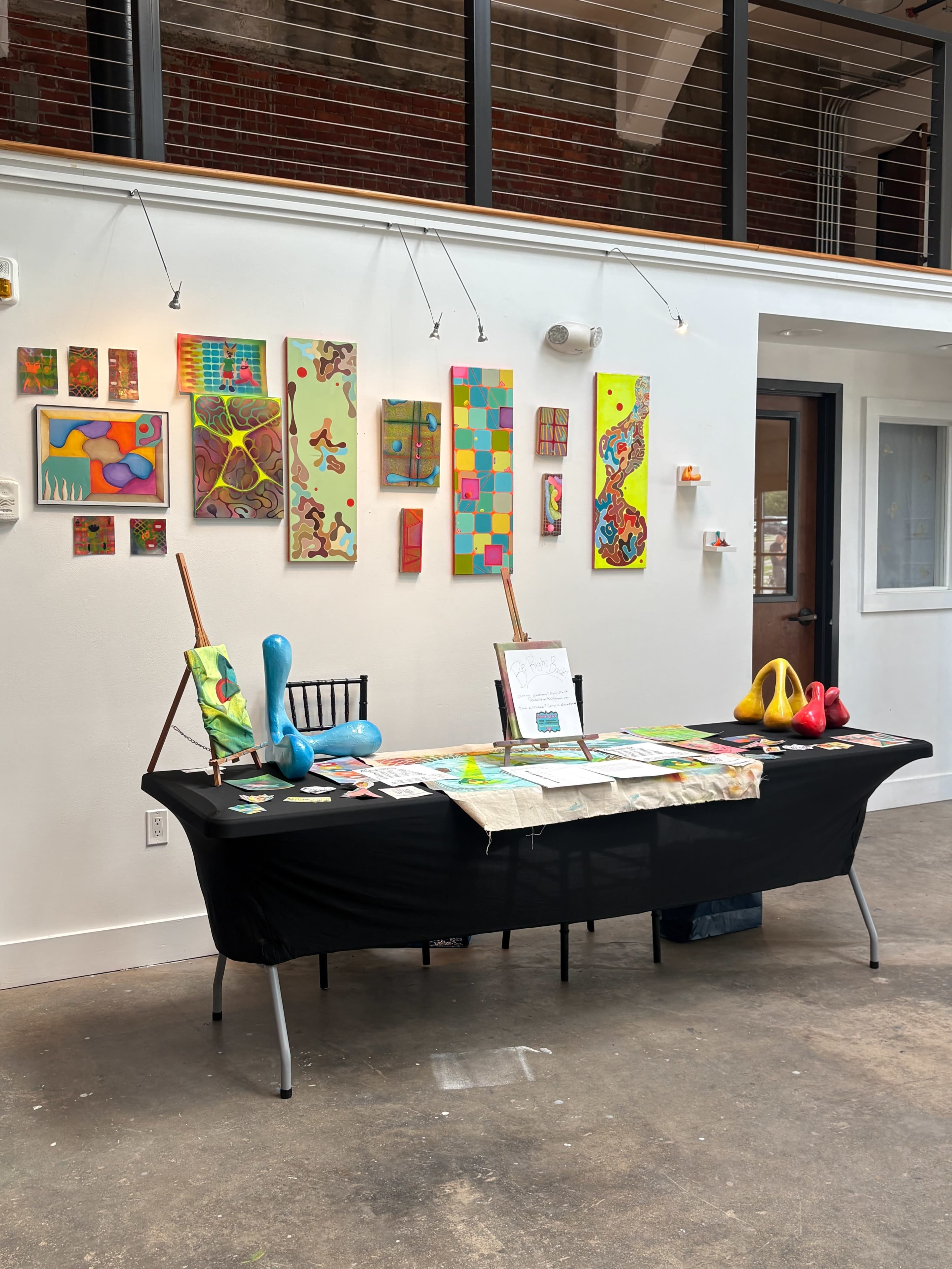 A black table is set up in an art gallery, displaying colorful artwork and sculptures against a backdrop of vibrant paintings on the wall.