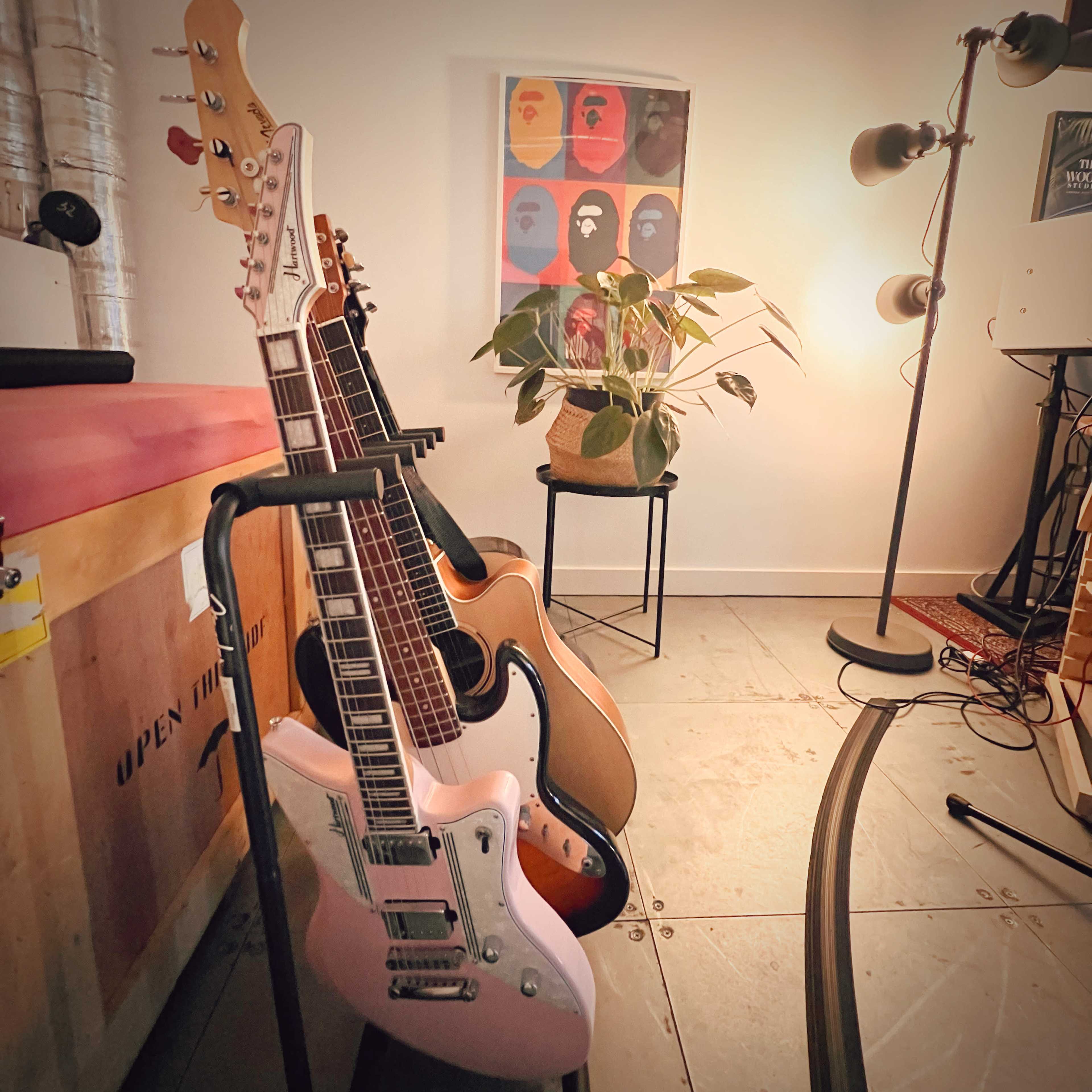 A collection of guitars on stands, a potted plant, a lamp, and a colorful framed poster in a well-lit room.
