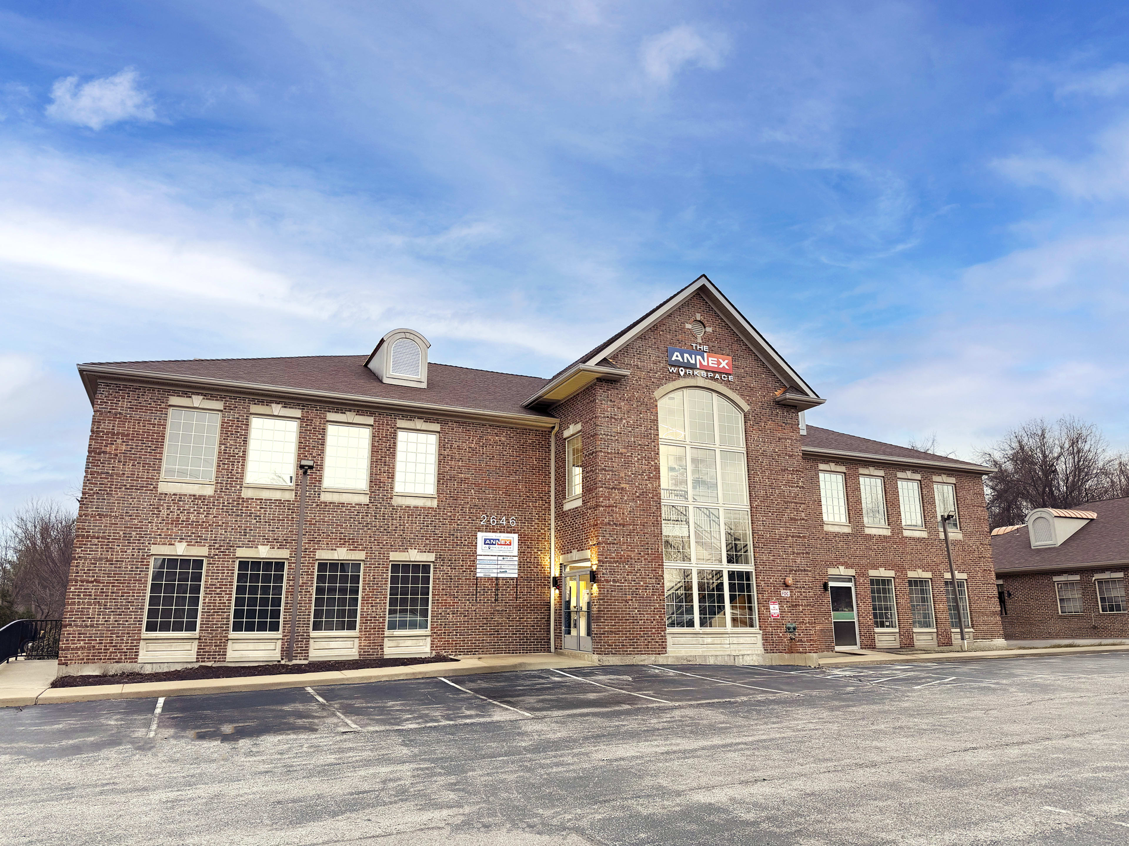 The image shows a two-story brick office building with large windows and a central entrance, surrounded by an asphalt parking lot.