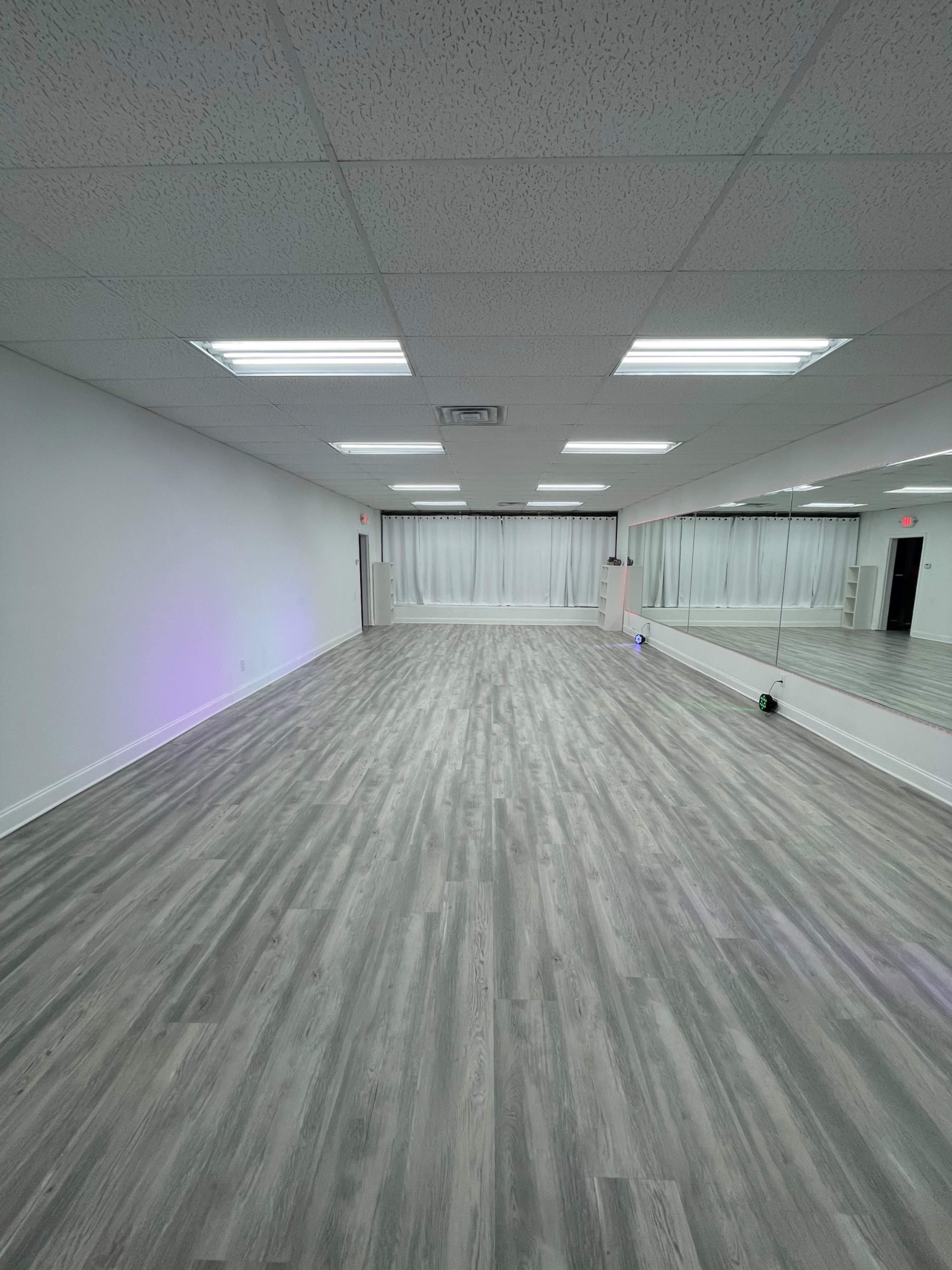 An empty room with mirrored walls, light-colored wood flooring, and fluorescent ceiling lights.