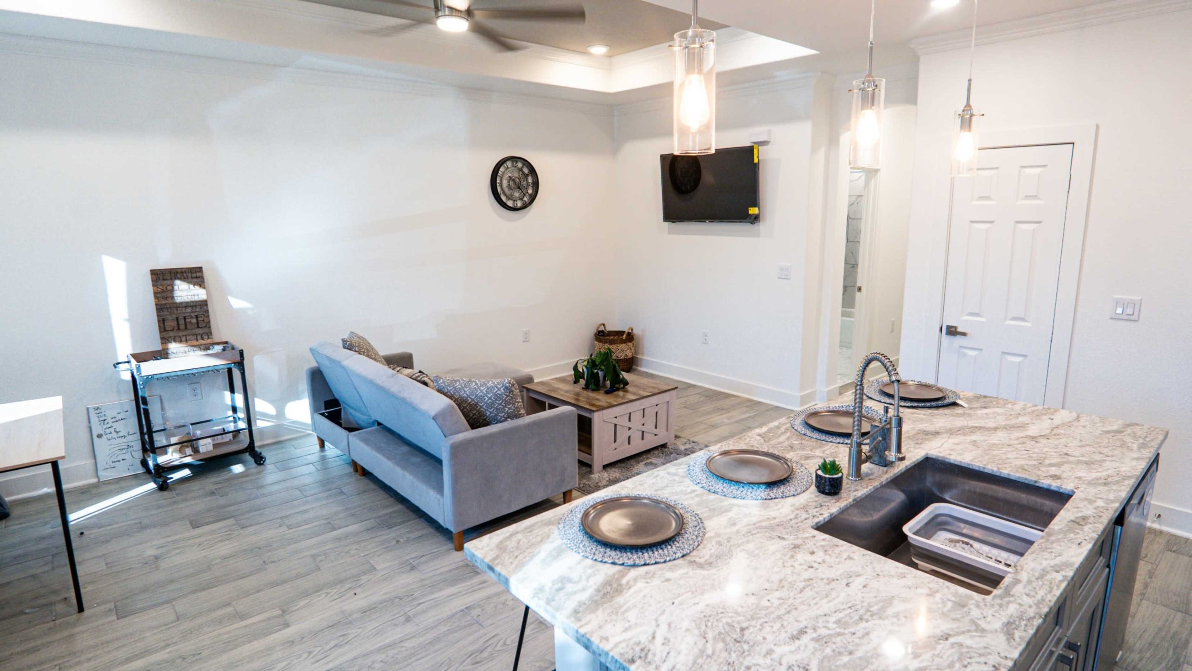 The image shows a modern living space featuring a gray couch, a coffee table, a kitchen island with a sink, and a wall-mounted television.