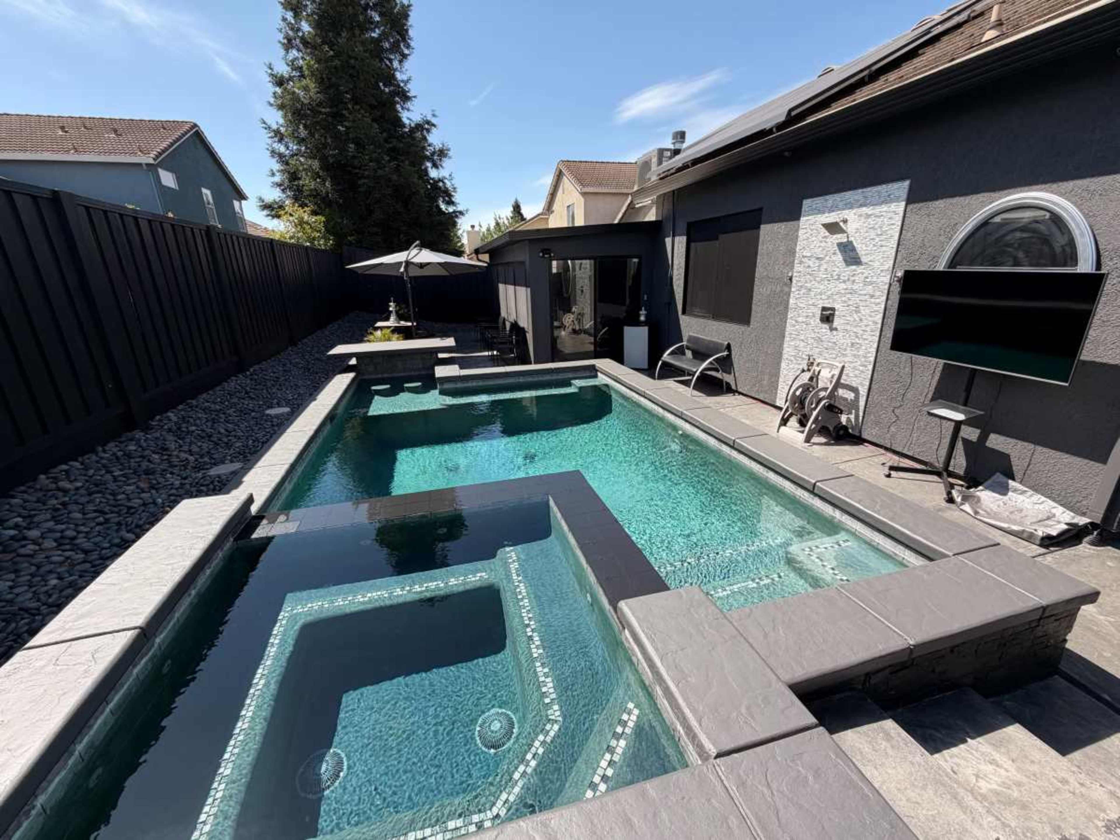 The image shows a modern backyard featuring a rectangular swimming pool surrounded by a stone deck, with patio furniture and a TV mounted on the wall nearby.