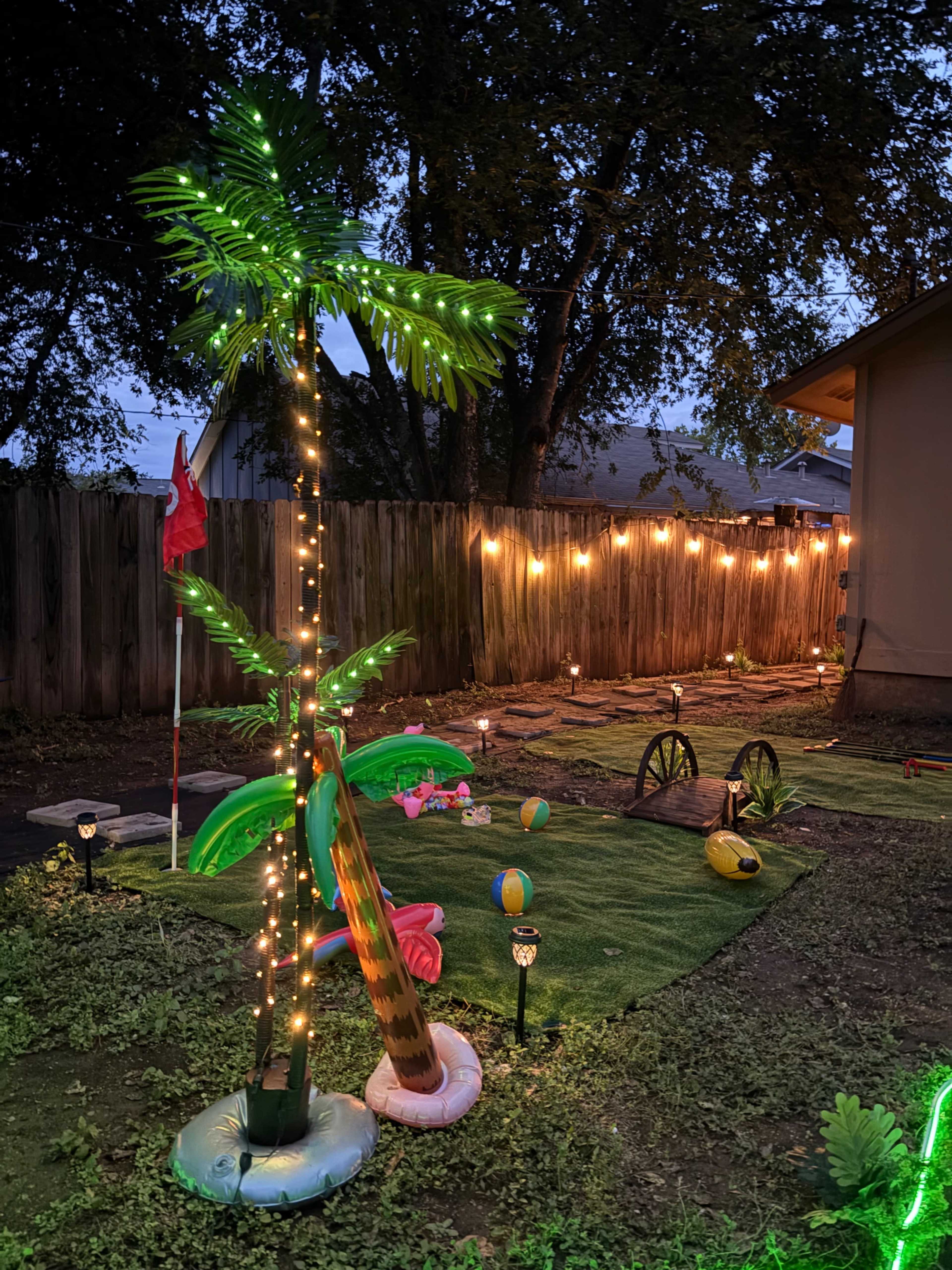 The image shows a backyard scene with a decorated palm tree illuminated by lights, alongside various colorful decorations and a wooden fence in the background.