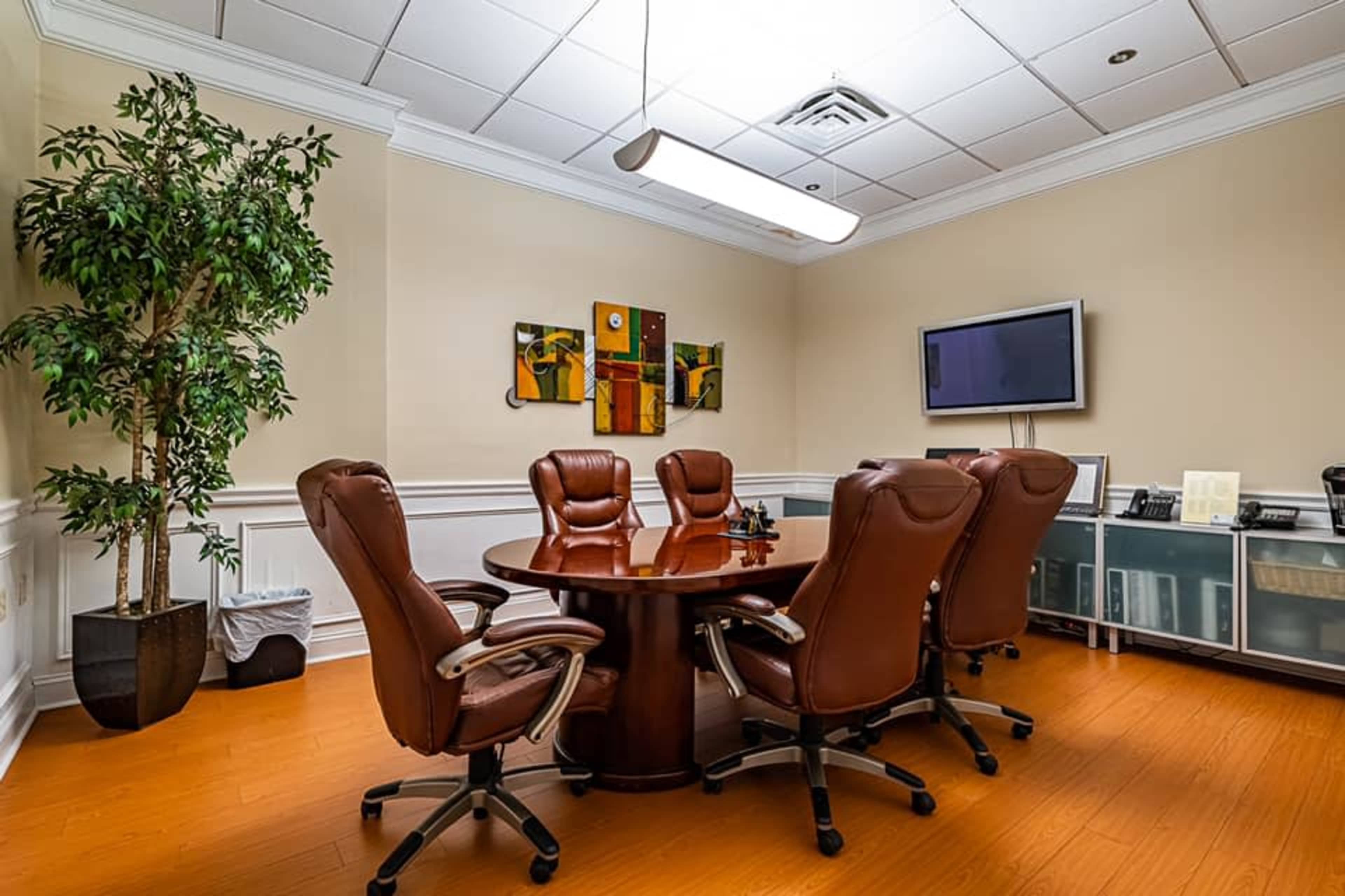 A conference room features a round wooden table surrounded by six brown leather chairs, with a potted plant and artwork on the walls.