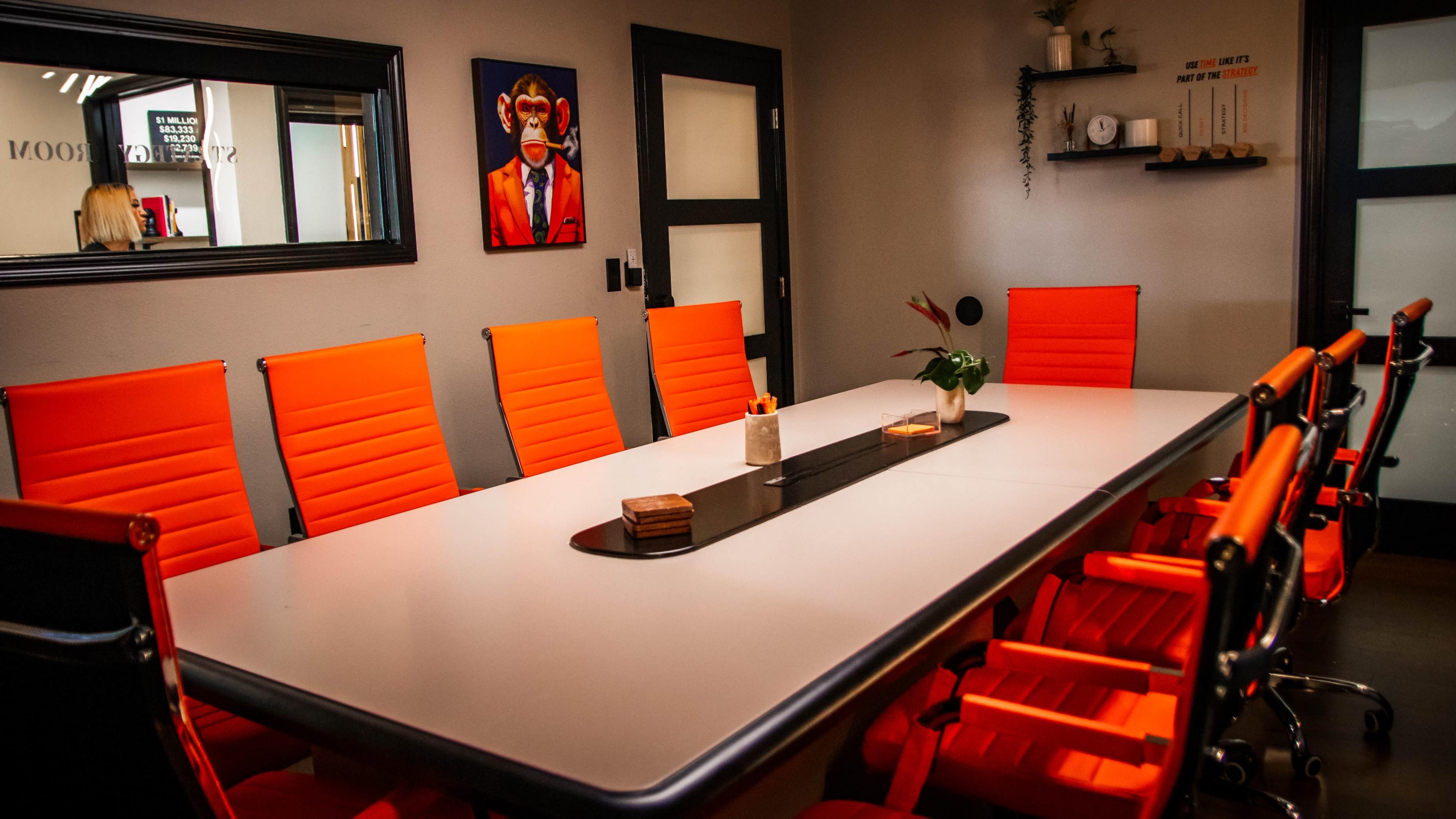 The image shows a modern conference room with a long white table surrounded by bright orange chairs, and a wall featuring a mirror and artwork.
