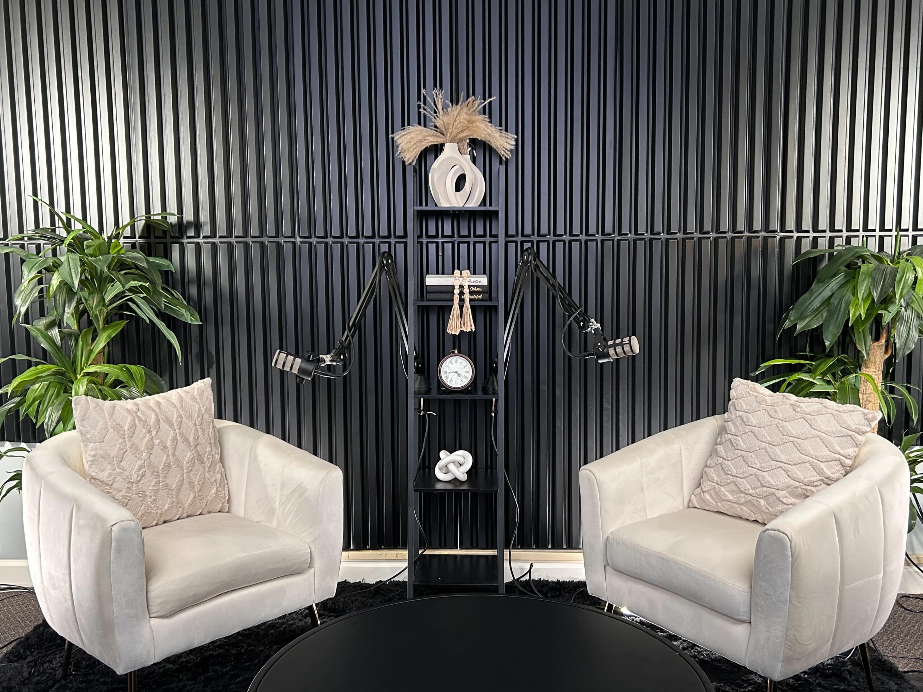 The image depicts a modern podcast studio setup with two white armchairs, a round black coffee table, a shelf displaying decorative items, and audio equipment mounted on either side against a textured black wall.