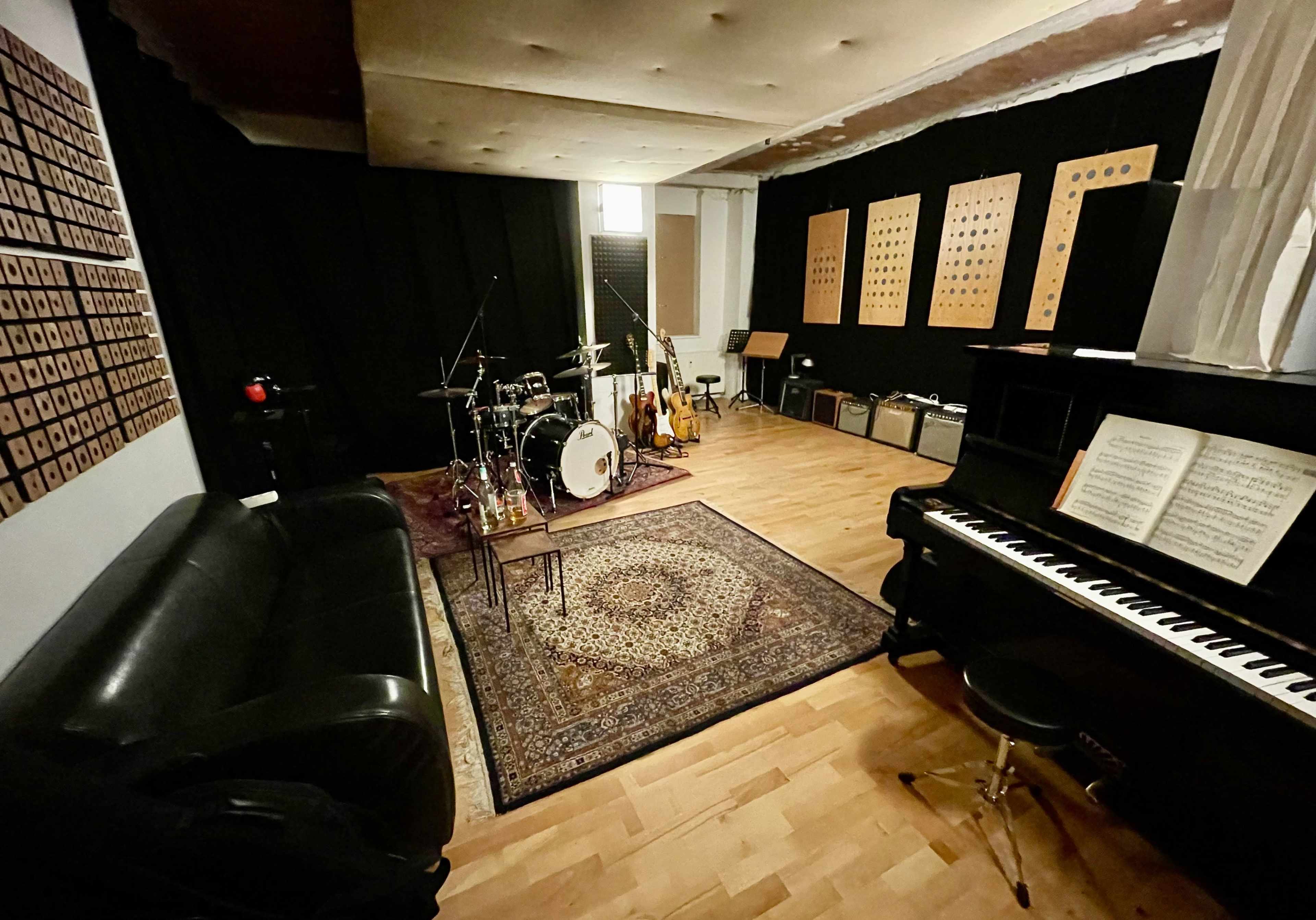 A music studio featuring a drum set, electric guitars, amplifiers, a grand piano, and a seating area with a black couch.