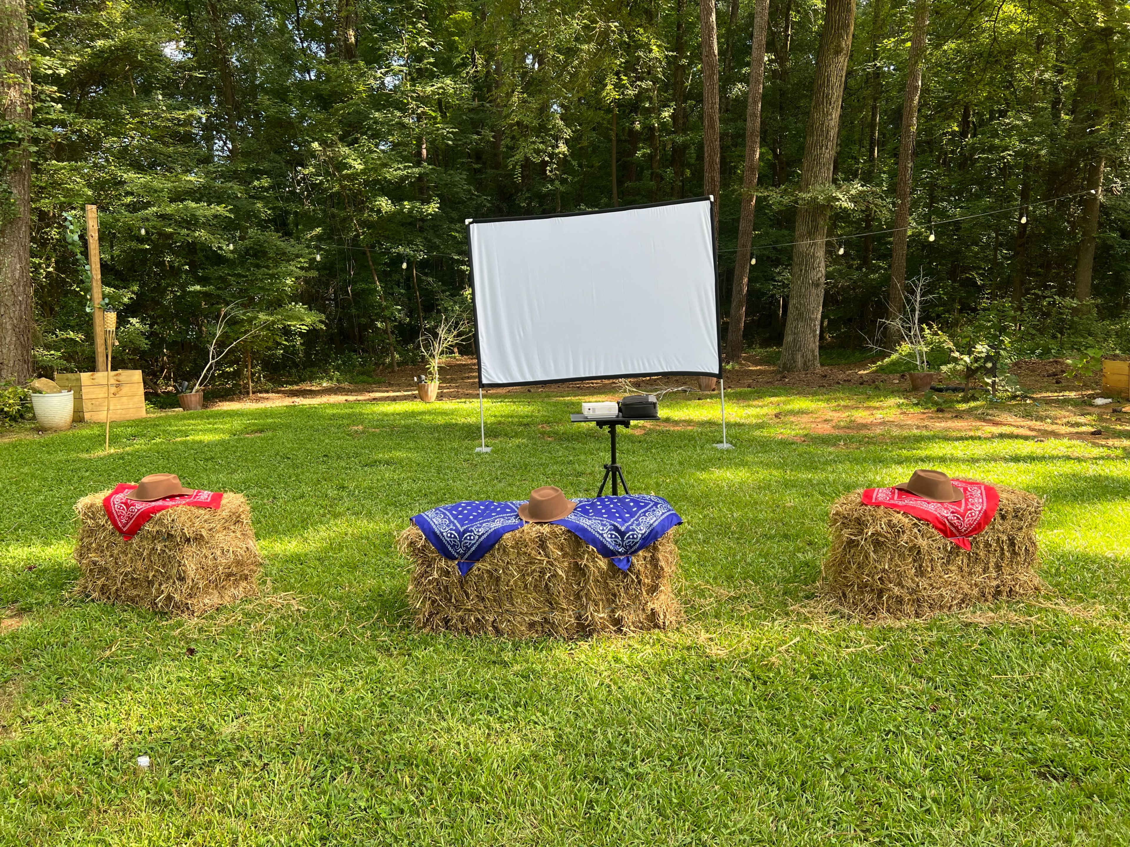 Three hay bales are arranged in a circle on grass, each topped with a cowboy hat and a colored bandana, facing a blank white projection screen.