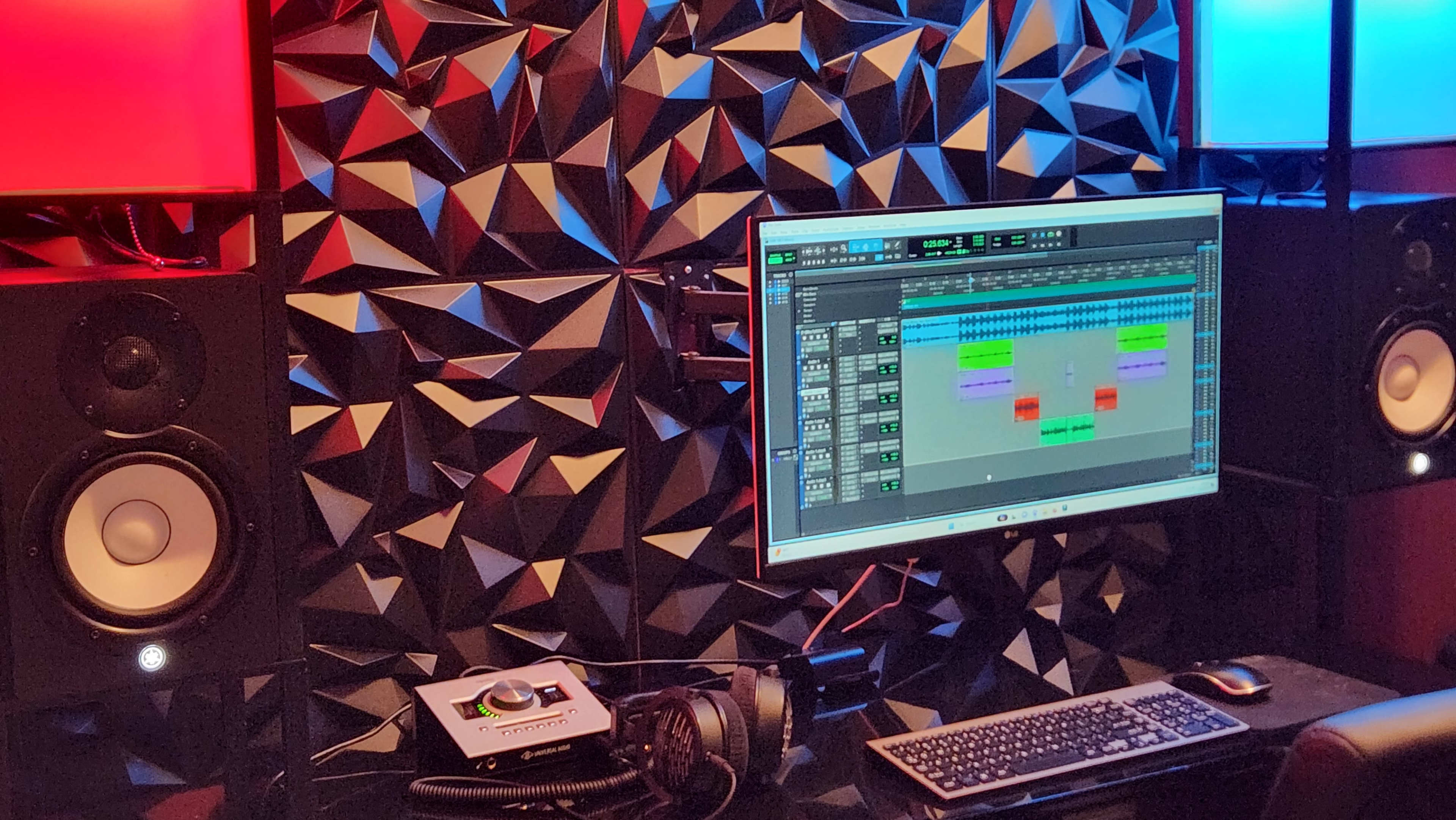 A sound studio setup featuring a computer monitor displaying a music production software, flanked by two large speakers, against a geometric patterned wall illuminated by red and blue lights.