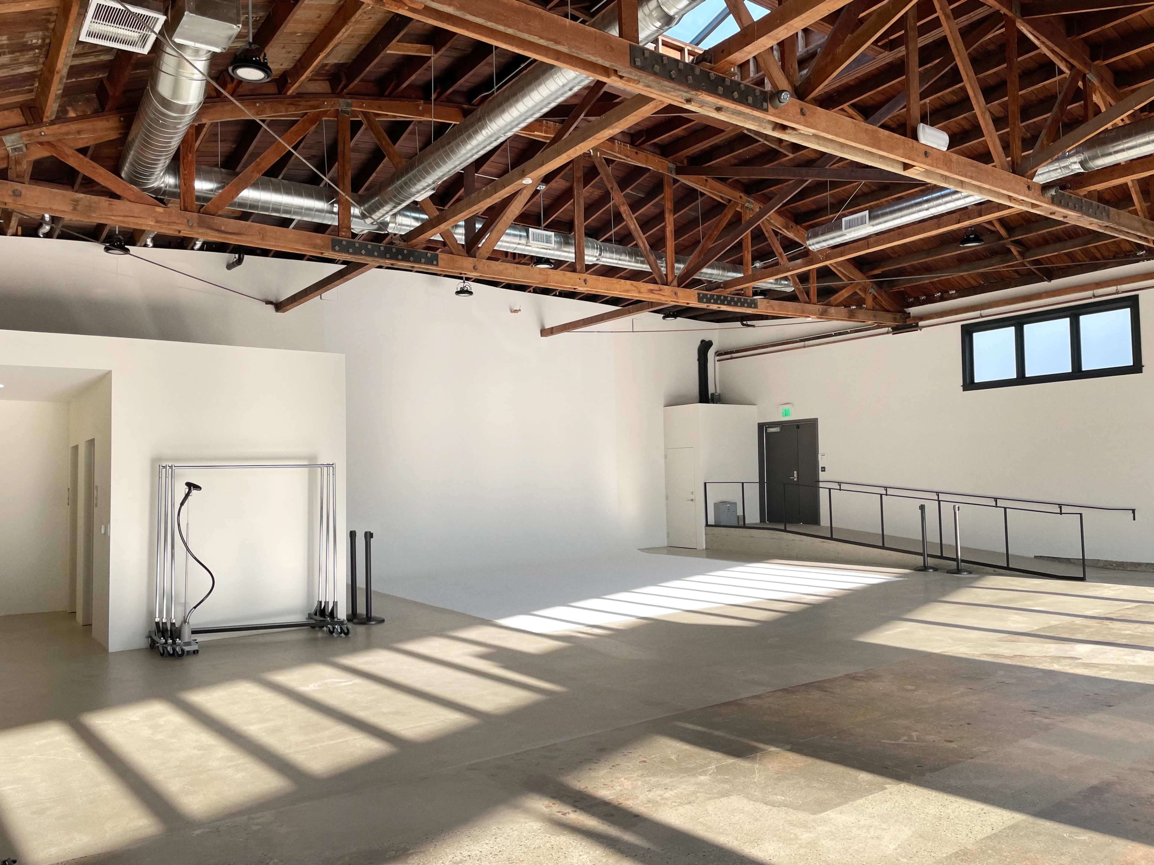 The image shows an industrial-style open space with high wooden ceilings, large windows, and minimal furniture, featuring a clean, empty area with natural light streaming in.