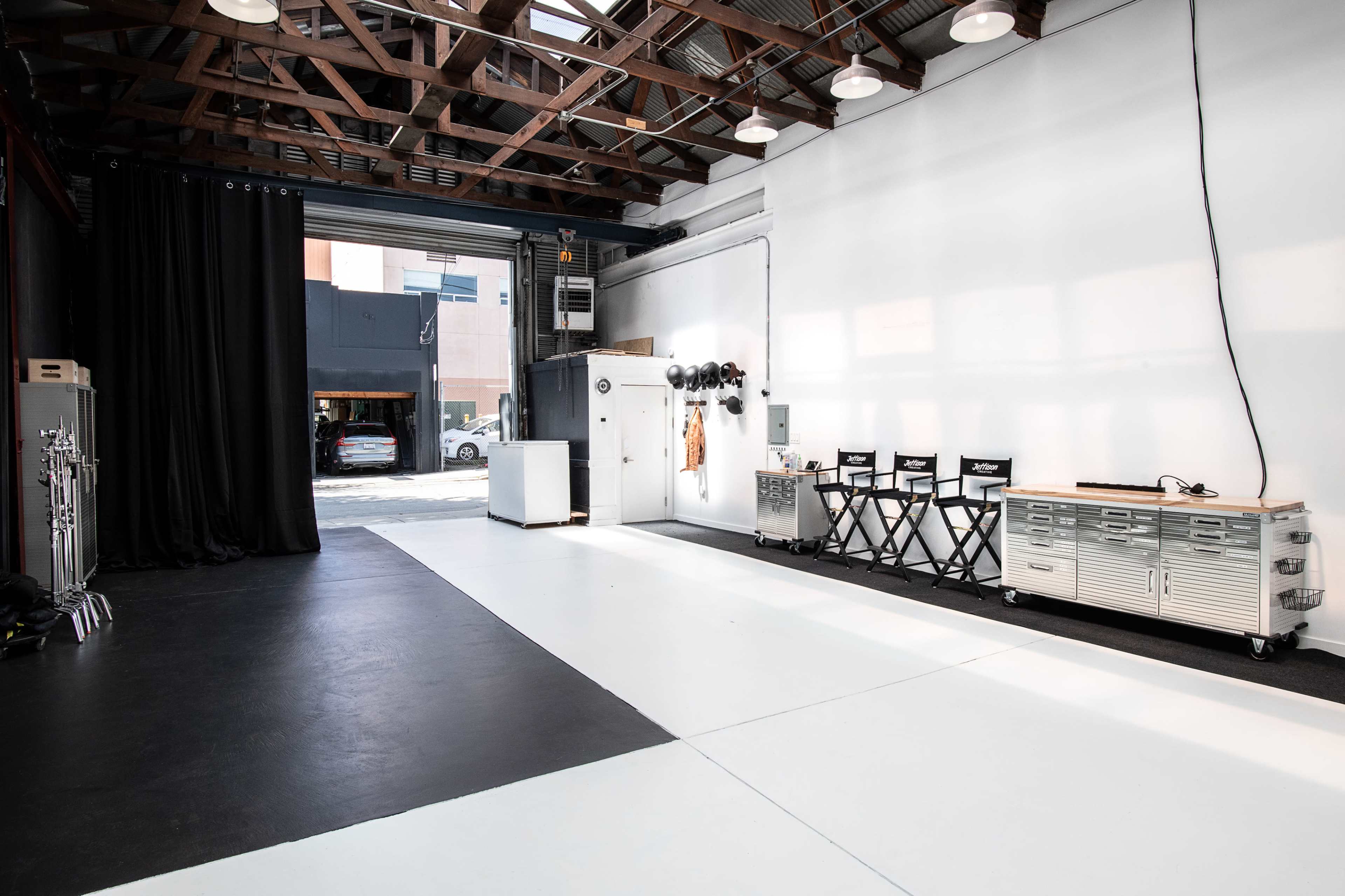 A spacious studio with black and white flooring, equipped with chairs, a storage unit, and large windows allowing natural light to enter.