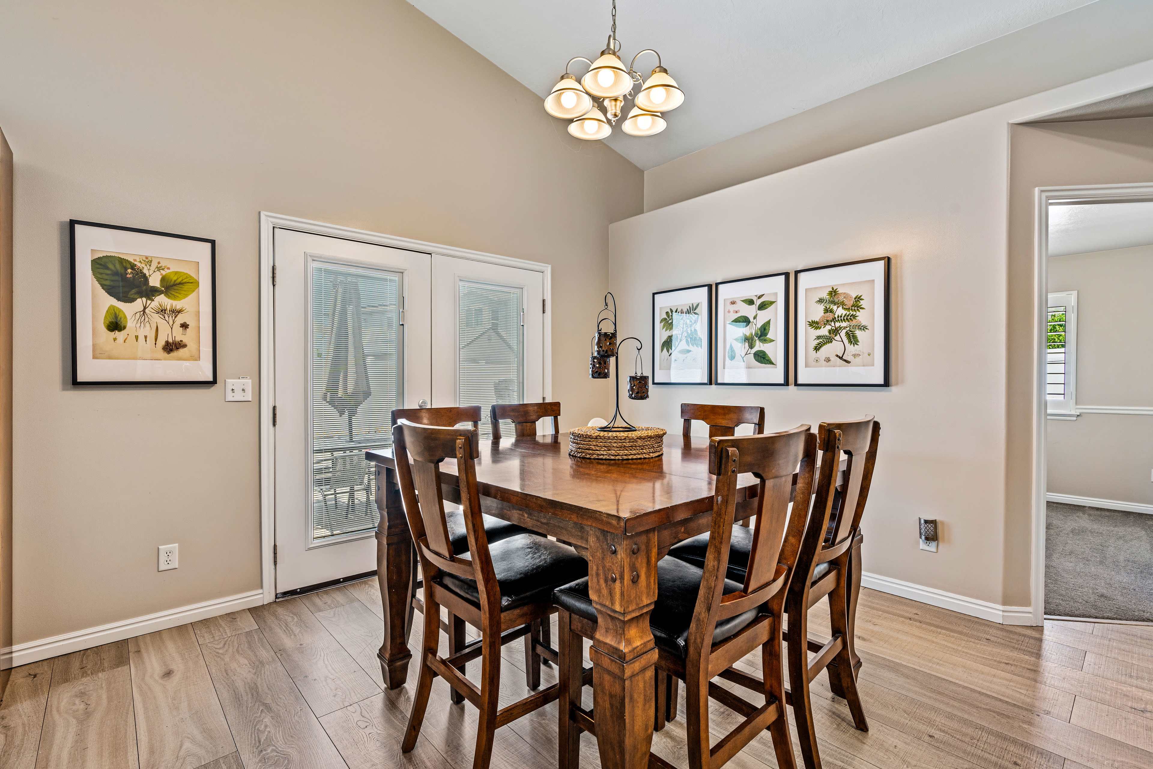 A wooden dining table surrounded by high-backed chairs is positioned in a well-lit dining area, with framed botanical prints on the walls and a chandelier overhead.