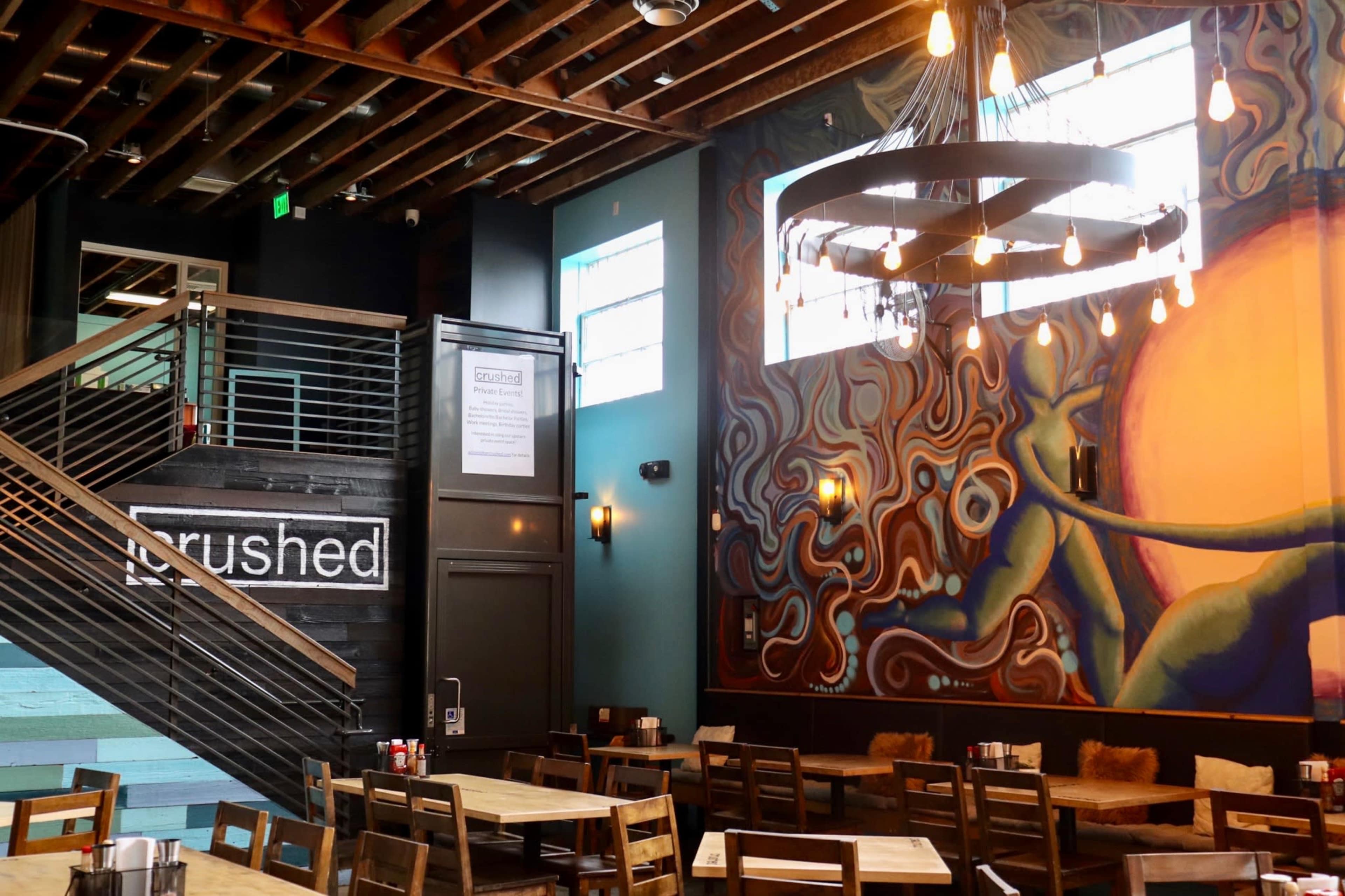 A spacious restaurant interior featuring wooden tables, a large mural on the wall, and a staircase leading to an upper level.