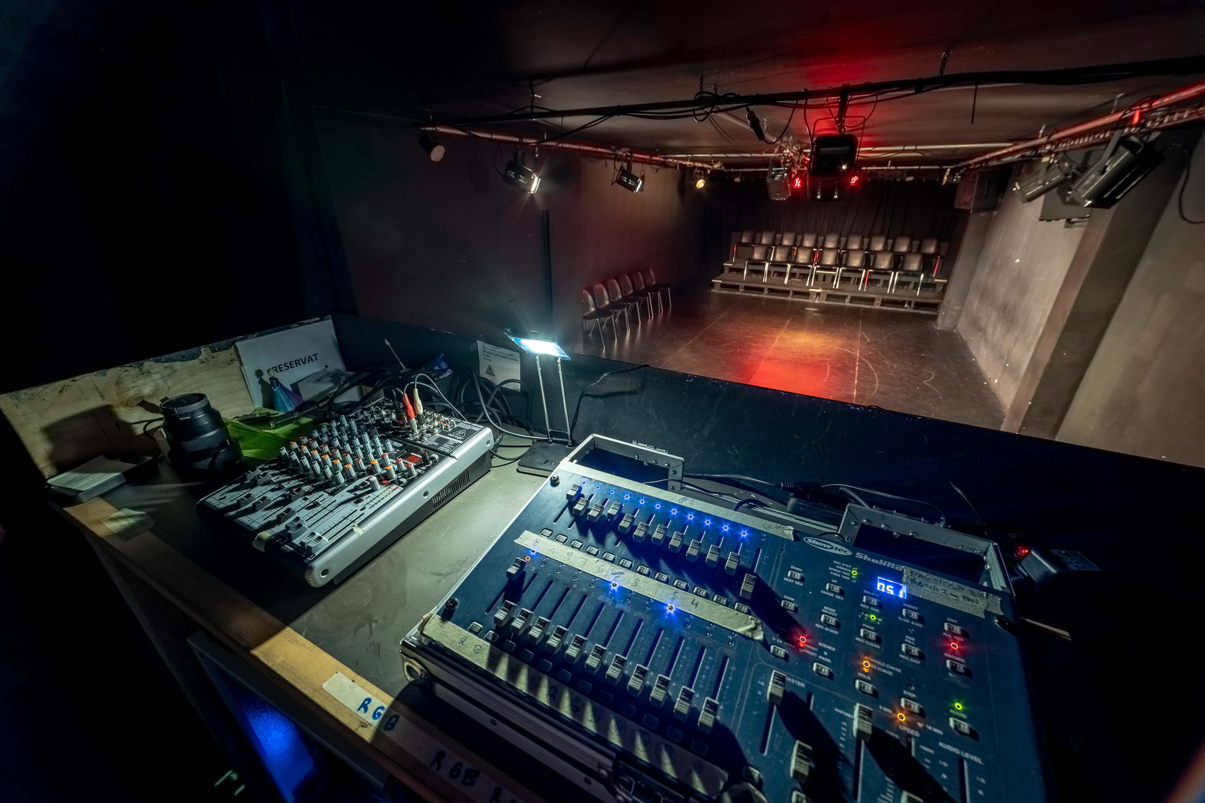A sound and lighting control panel overlooking an empty stage with rows of chairs arranged in the background.