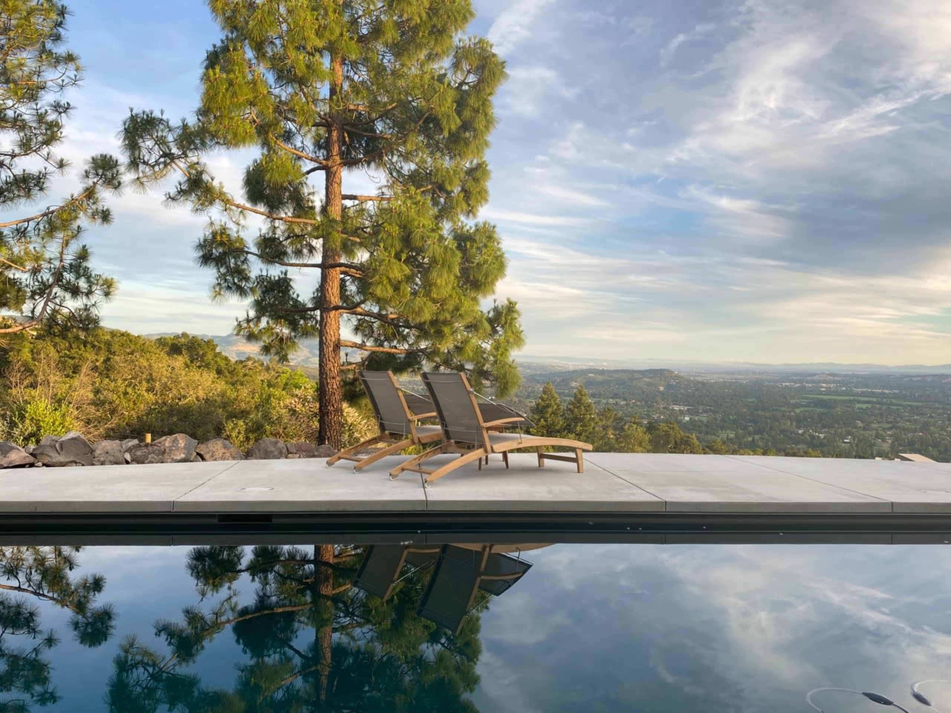 A swimming pool with a reflective surface sits next to three lounge chairs, set against a backdrop of trees and a distant landscape.