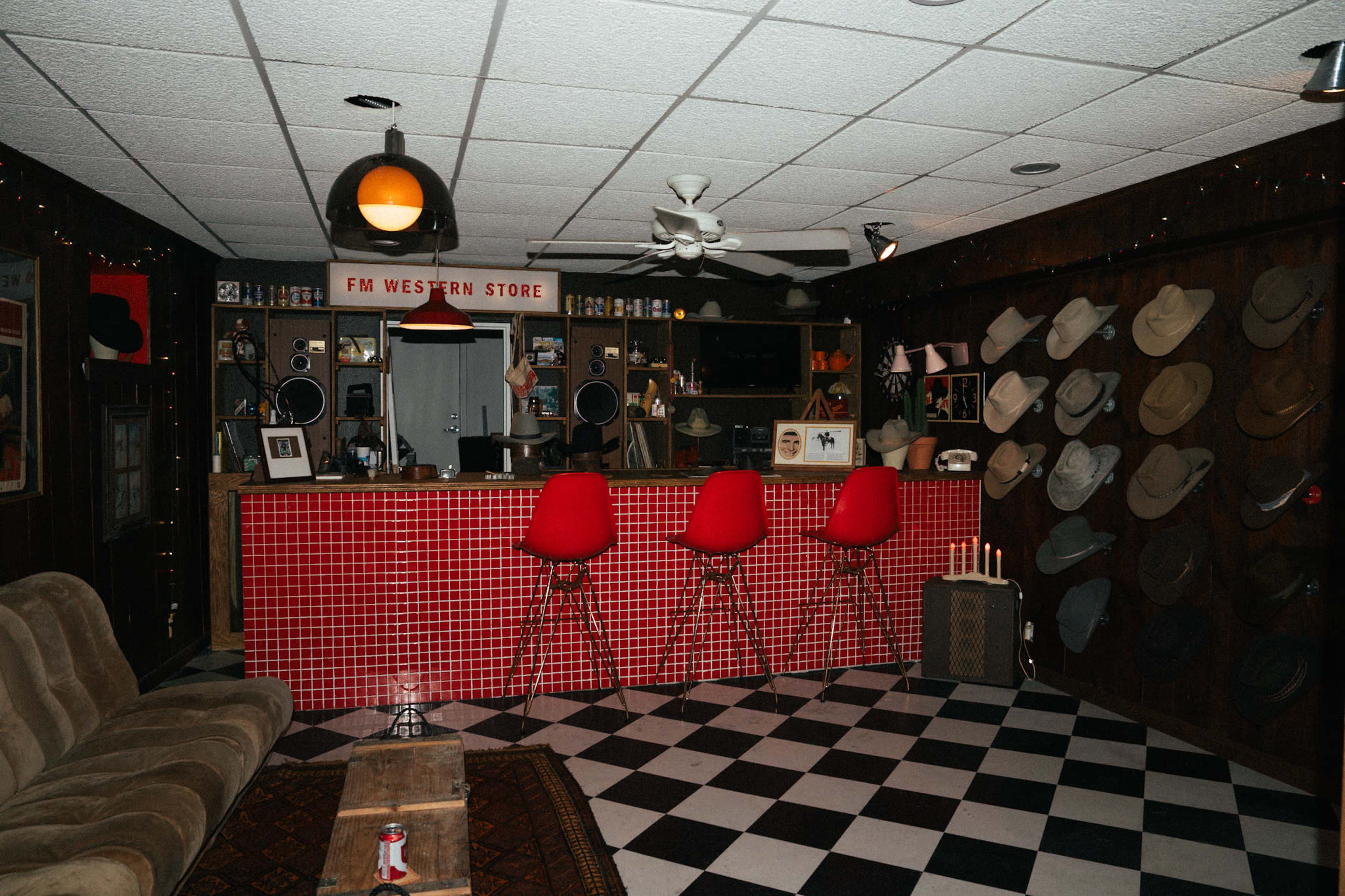 The image shows a room designed as a bar or store with a tiled counter, red bar stools, a wall lined with hats, and a checkered floor.