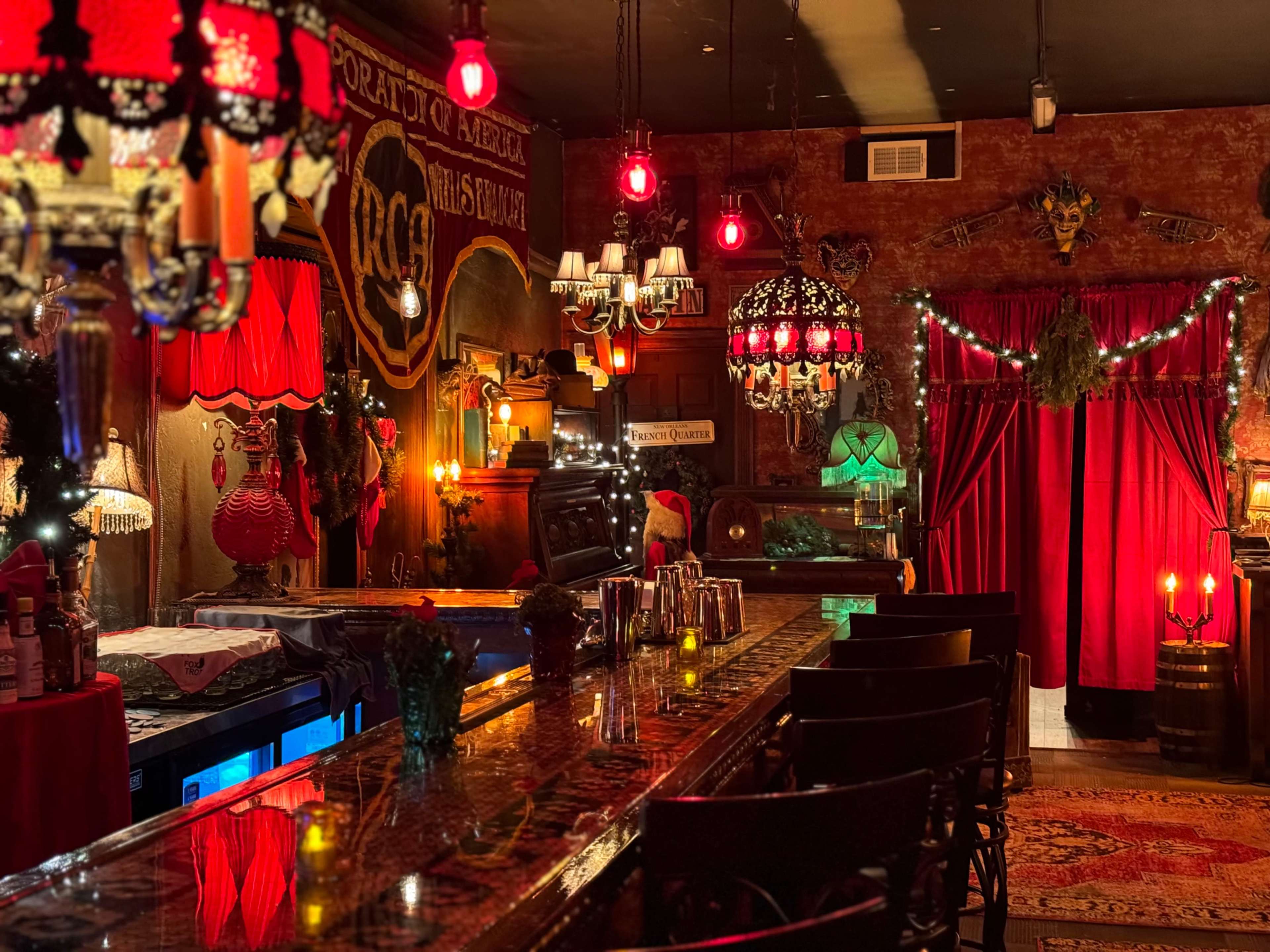 A dimly lit bar with ornate decorations, featuring a long bar counter, various hanging lights, and red curtains.