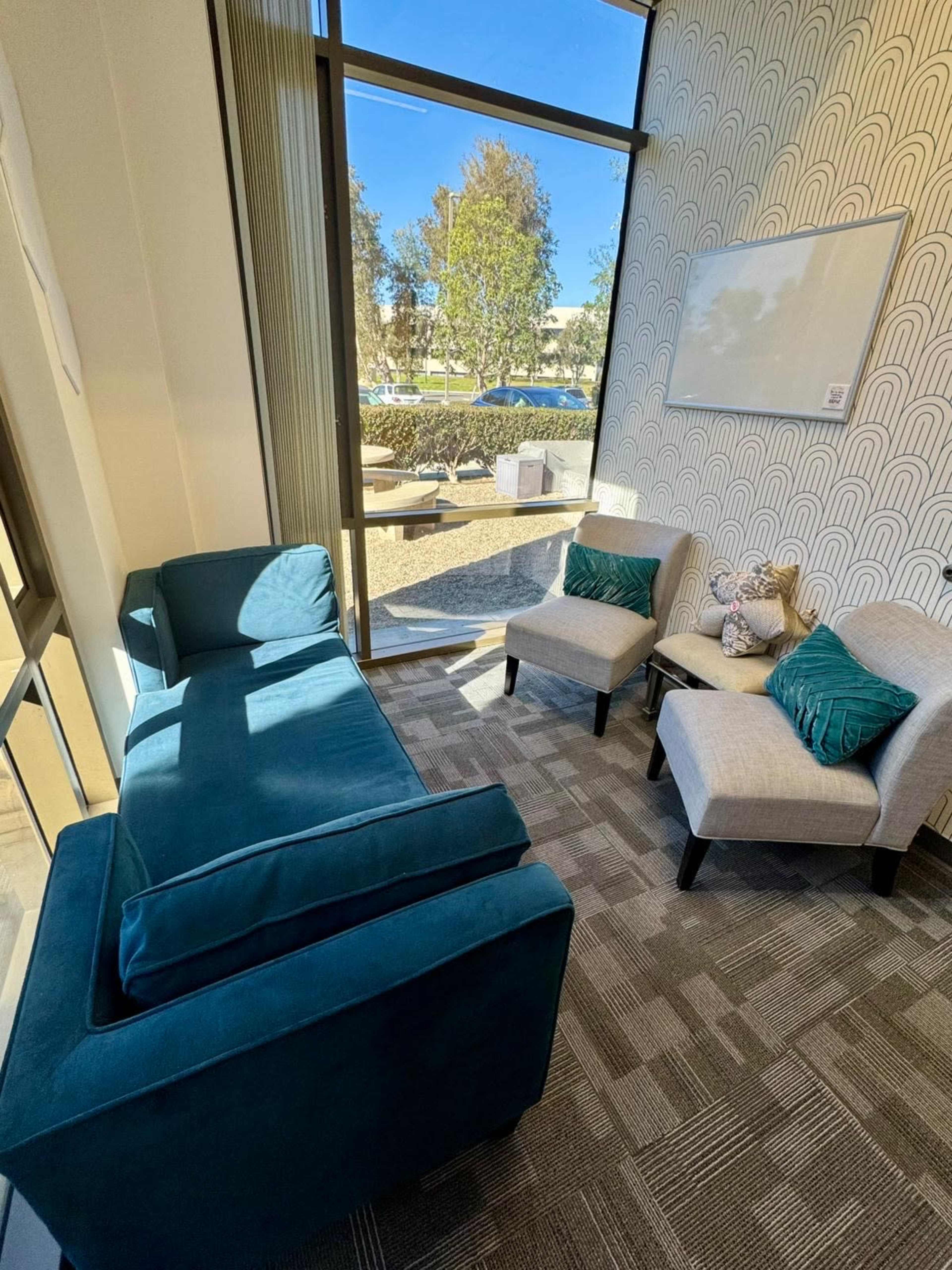 The image shows a cozy seating area with a teal couch and two light-colored chairs positioned near a large window, overlooking a landscaped outdoor space.